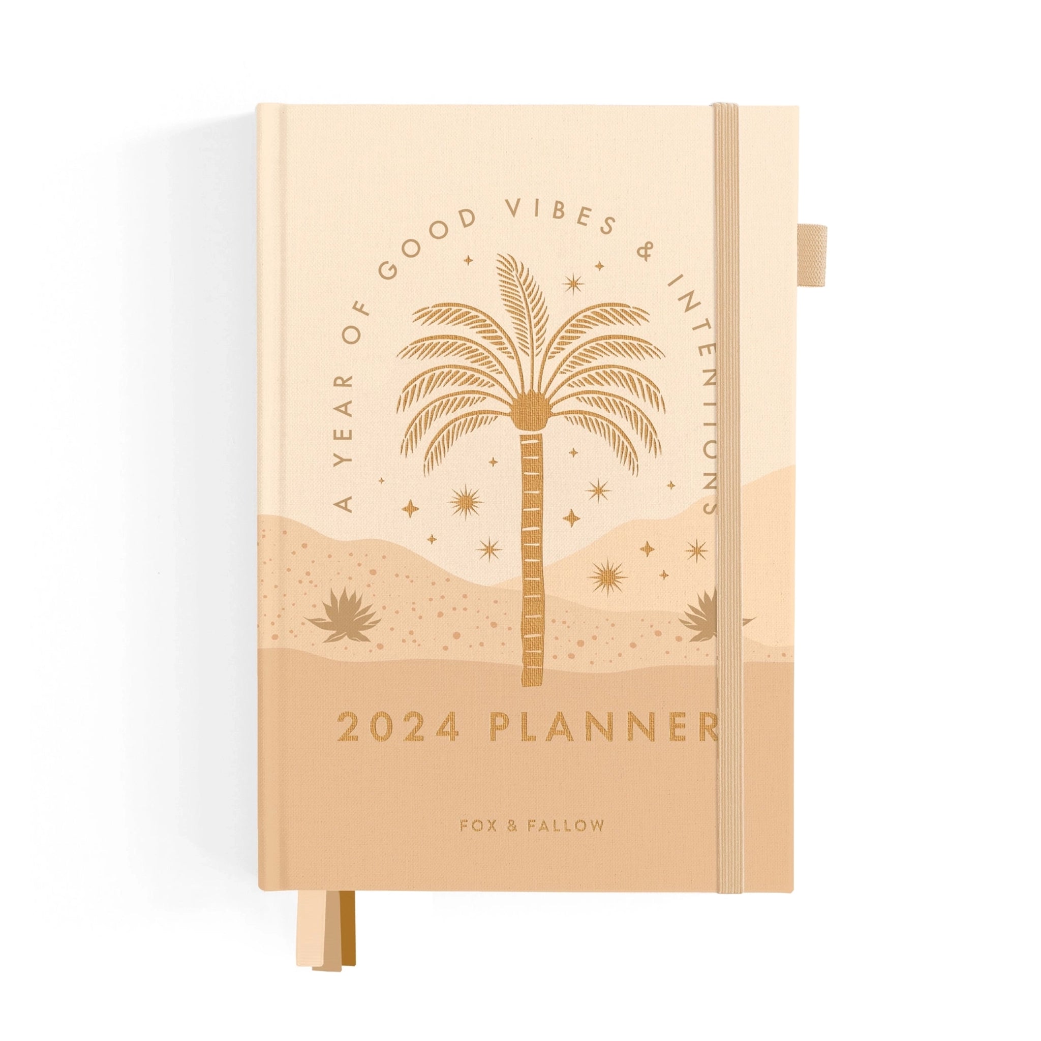 On a white background is a tan planner with gold foiled &quot;2024 Planner&quot; text in the center as well as arched text over a palm tree design that reads, &quot;A Year Of Good Vibes &amp; Intentions&quot; and a light pink elastic loop for keeping shut.