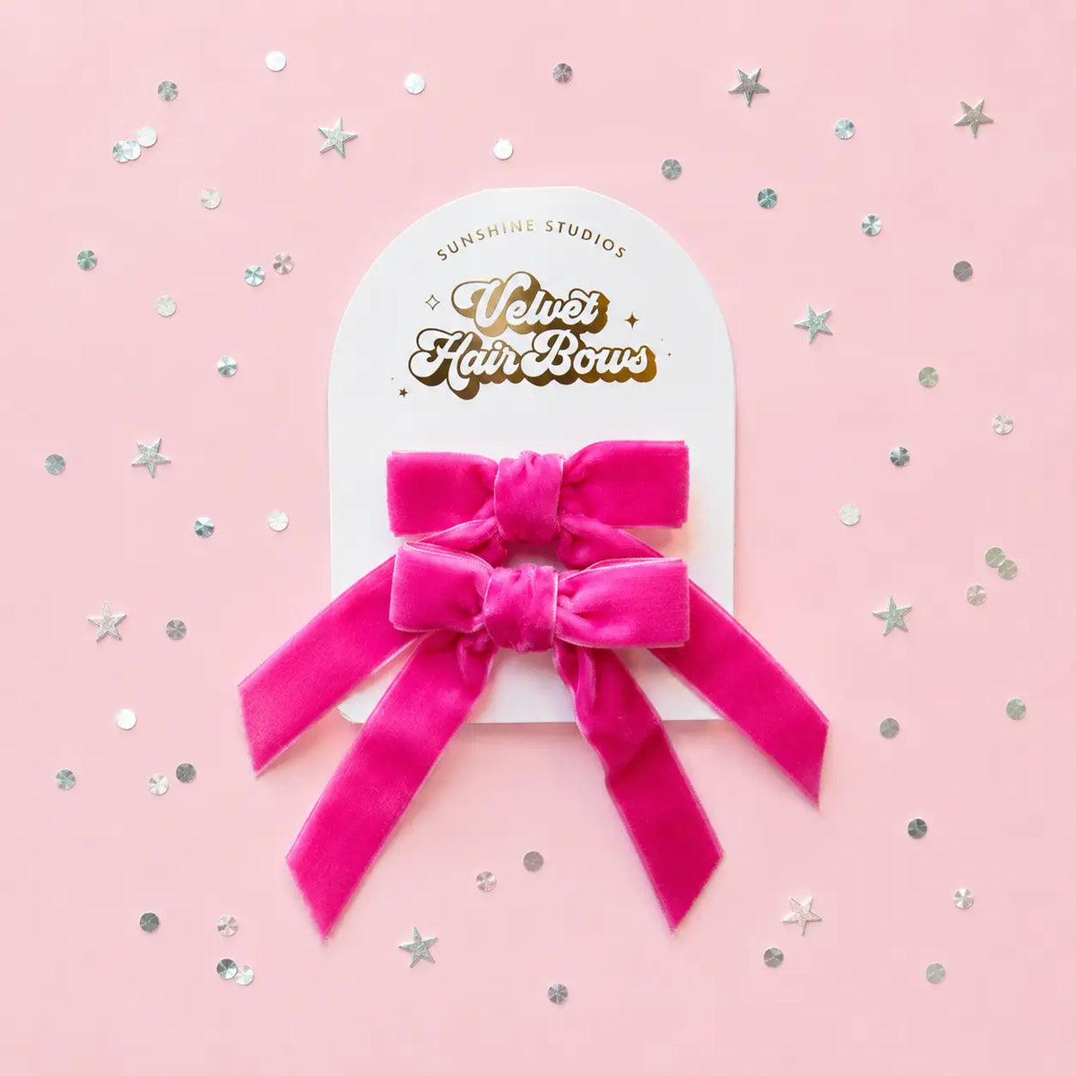 A bright pink velvet hair bow attached to a white display card reading 'Sunshine Studios Velvet Hair Bow,' photographed on a light pink background with scattered silver star and dot confetti.