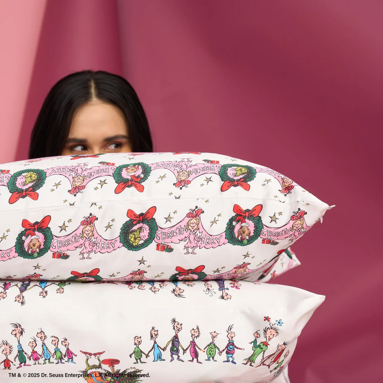 A person peeks over a stack of two white satin pillows; the top pillowcase features repeating Grinch holiday illustrations with wreaths and red bows, while the bottom shows a line of Whoville characters, set against a deep pink backdrop.