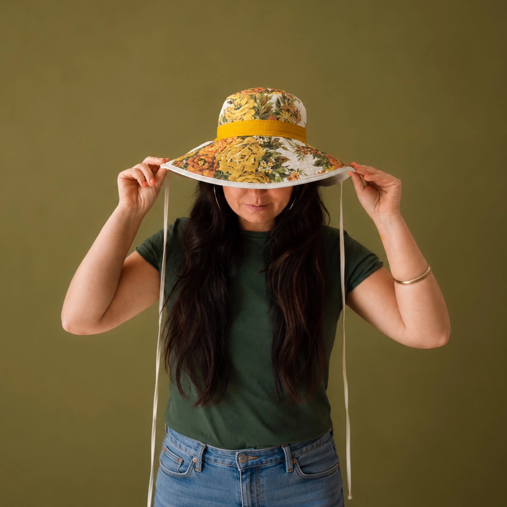 A wide-brim sun hat with a white base and a dense yellow, orange, and green floral print, featuring a mustard yellow band and white chin straps, set against a solid olive green background.