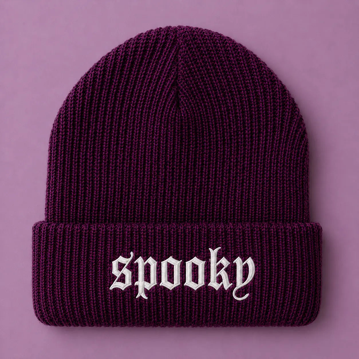 A dark purple ribbed knit beanie with the word 'spooky' embroidered in white Gothic lettering on the front cuff, set against a solid lavender background.