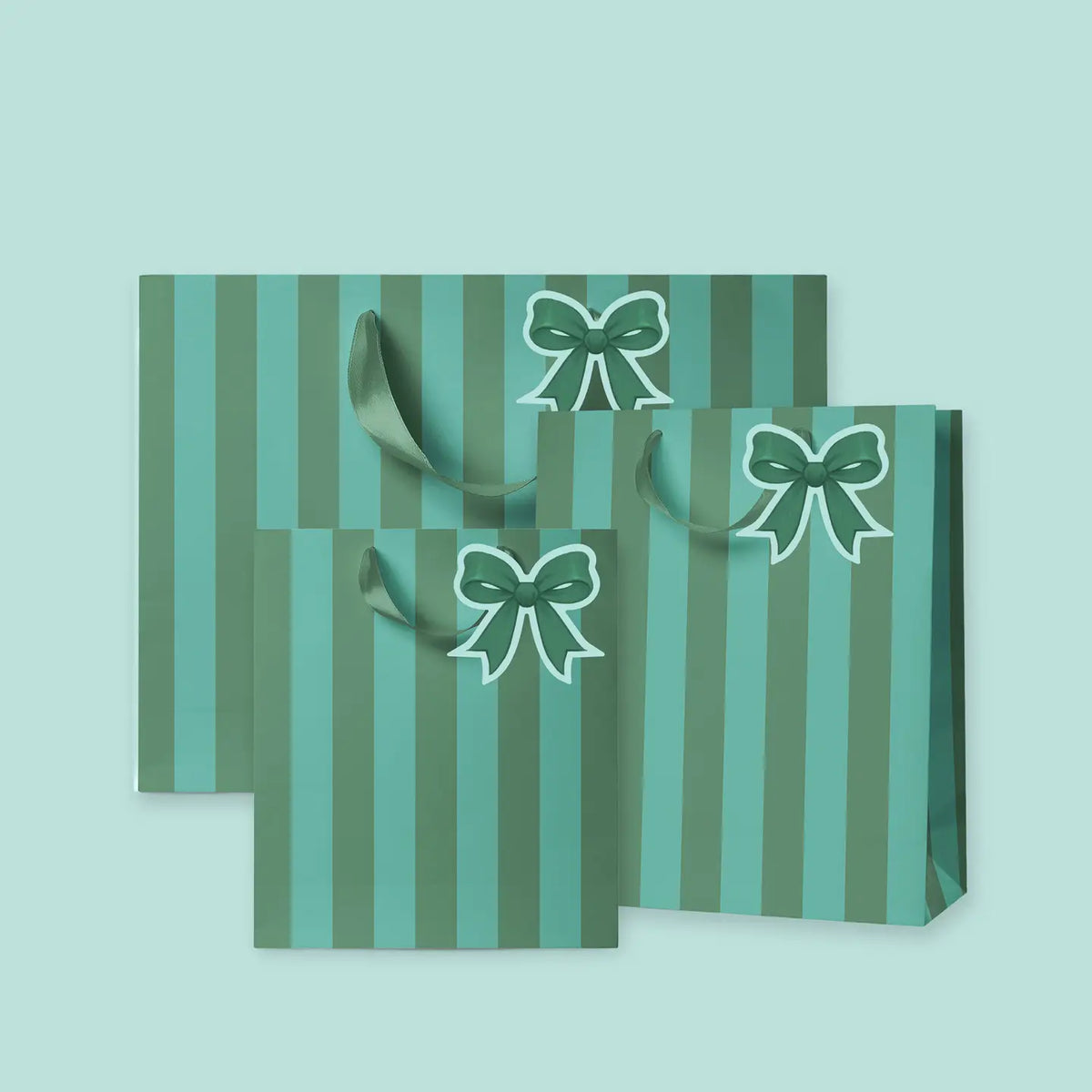 Three gift bags with vertical stripes in shades of green and teal, featuring printed green bows with white outlines and matching green handles, set against a solid pale mint green background.
