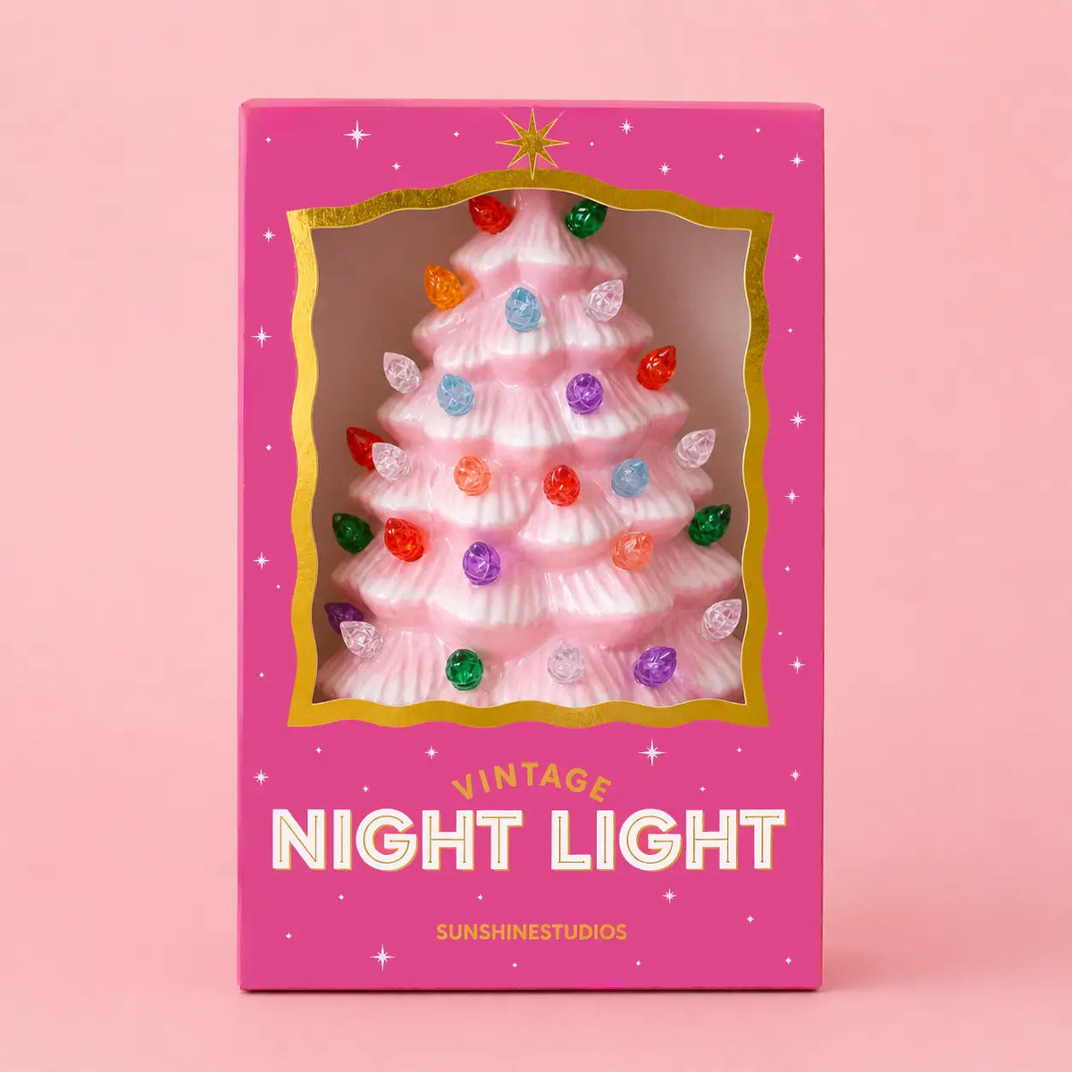 A pink Christmas tree-shaped night light in its original packaging, featuring a clear window that shows the light on, with colorful bulb accents. The box is pink with gold trim and white text reading 'VINTAGE NIGHT LIGHT' and 'SUNSHINESTUDIOS'.