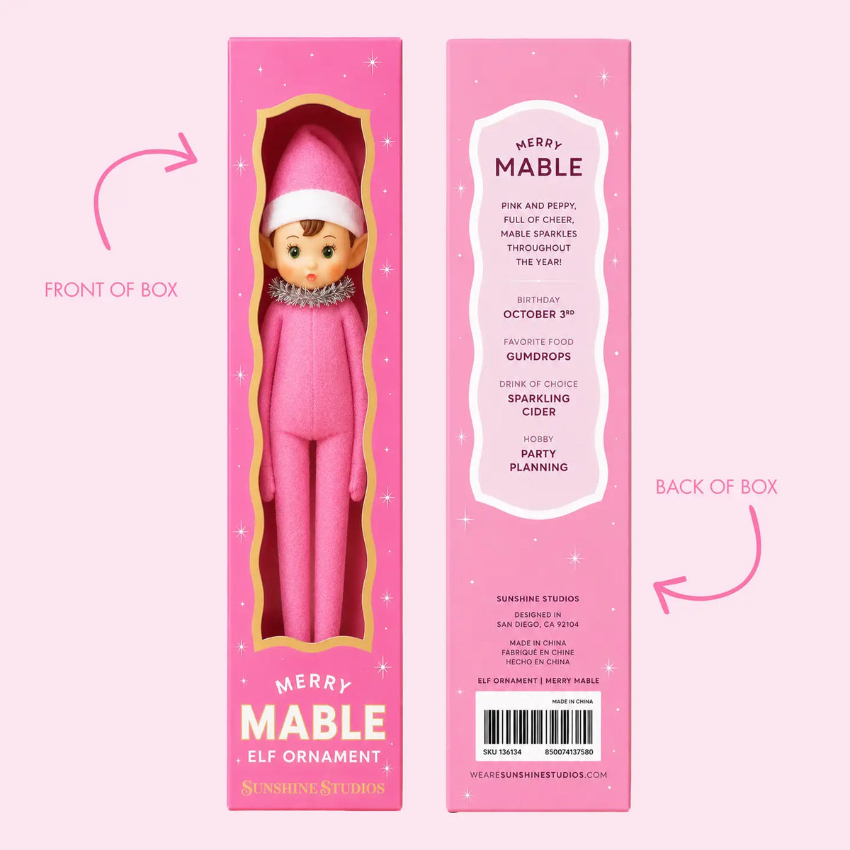 Front and back views of a pink Christmas elf ornament box labeled Merry Mable, with a doll visible through the windowed front panel and printed product details on the back, on a pale pink background with arrow callouts.
