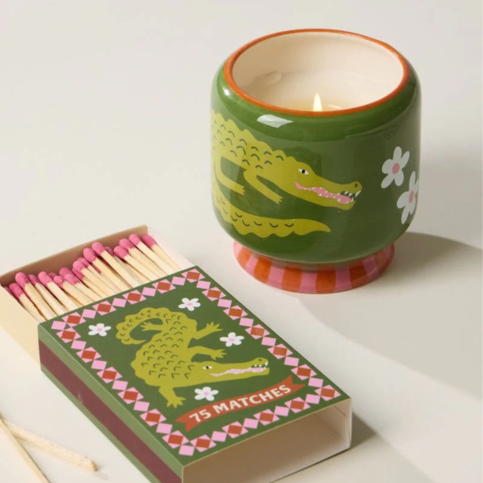 A green matchbox with a crocodile artwork on it and pink matches inside.  The box sits next to a candle that has a crocodile print on it.