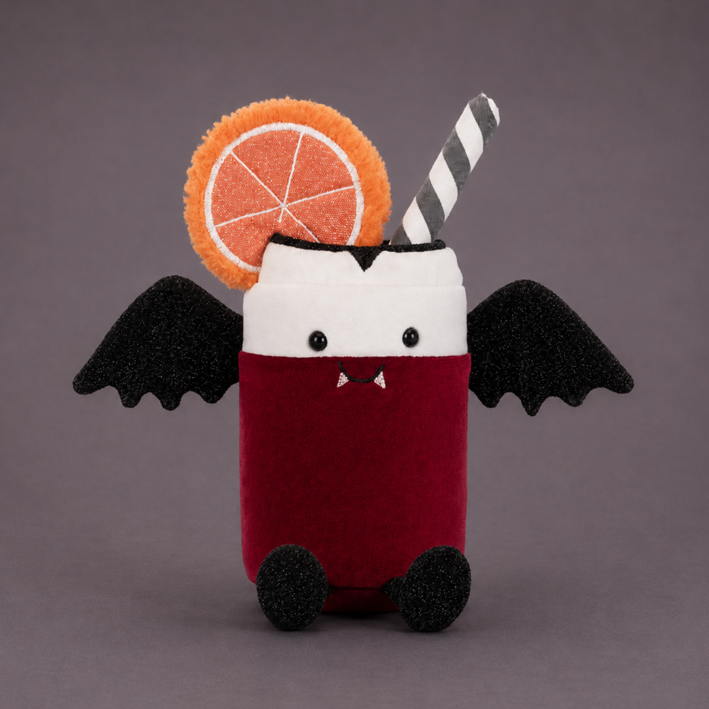 a red and white soda can shaped plush toy with a smiling stiched vampire face and black wings. 