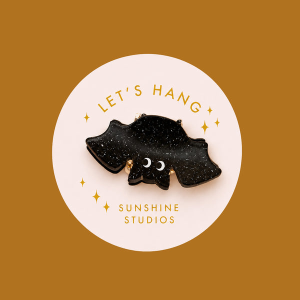 A black speckled bat shaped mini hair clip on a round piece of packaging that reads, 'Let's Hang Sunshine Studios'. 