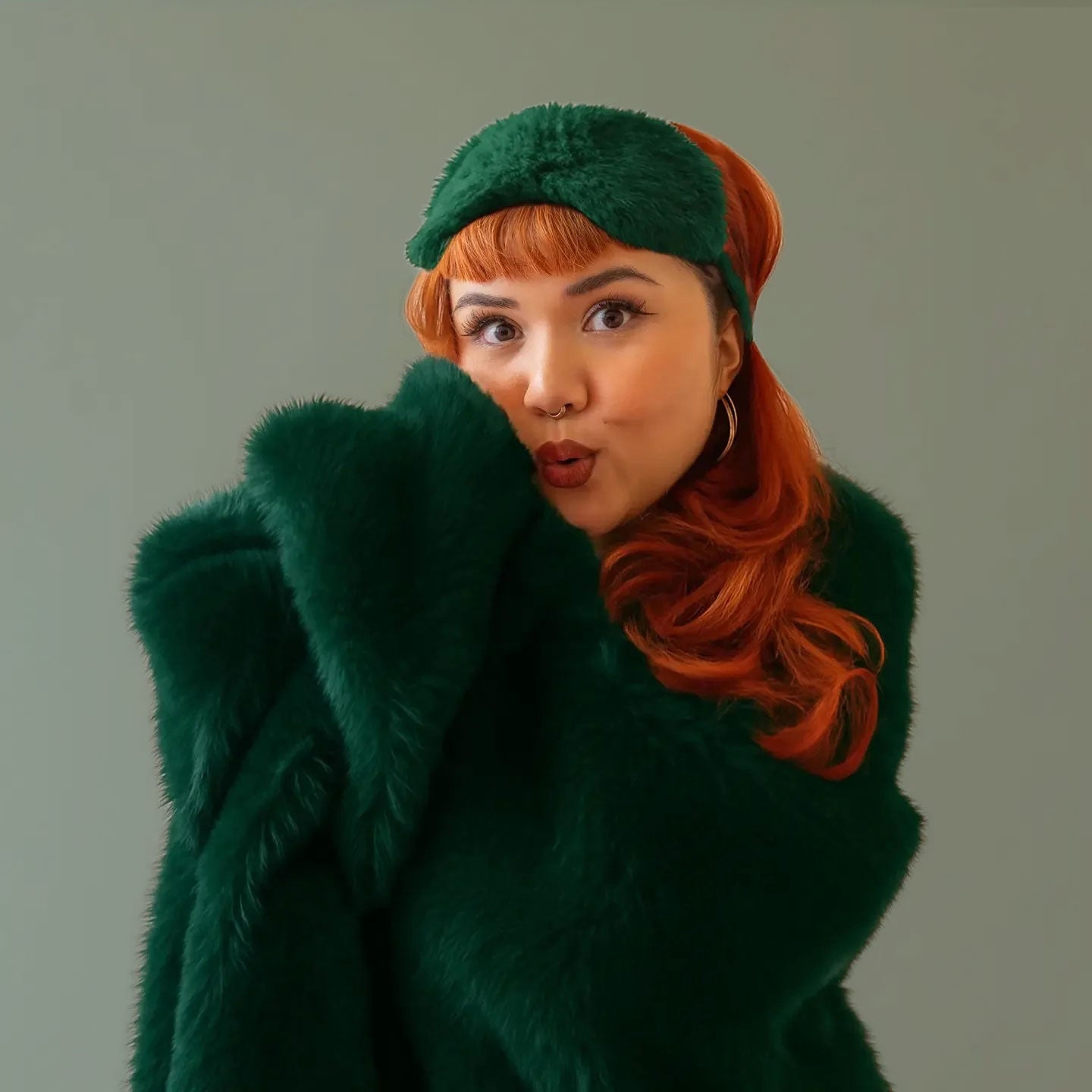 A red headed model wears a fuzzy emerald colored fur blanket with a green fur eye mask.