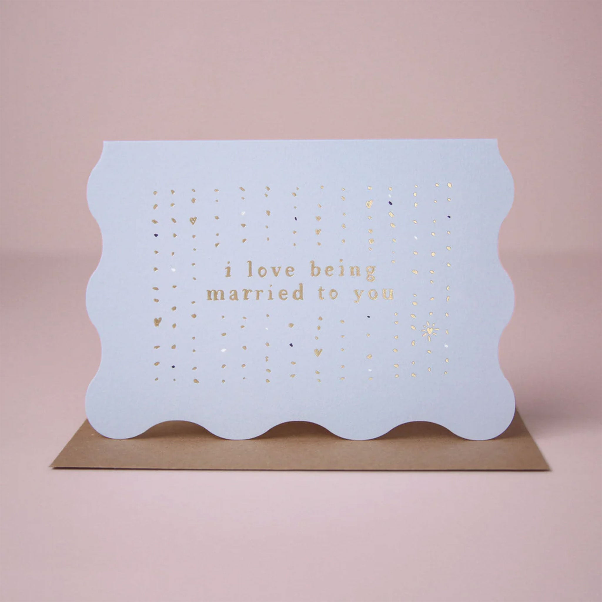 On a neutral background is a blue wavy edged card with gold foiled text that reads, &quot;i love being married to you&quot;.