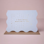 On a neutral background is a blue wavy edged card with gold foiled text that reads, "i love being married to you".