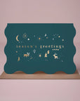 On a light pink background is a teal blue wavy edged greeting card with gold text and details that read, "season's greetings" along with a Kraft brown envelope.