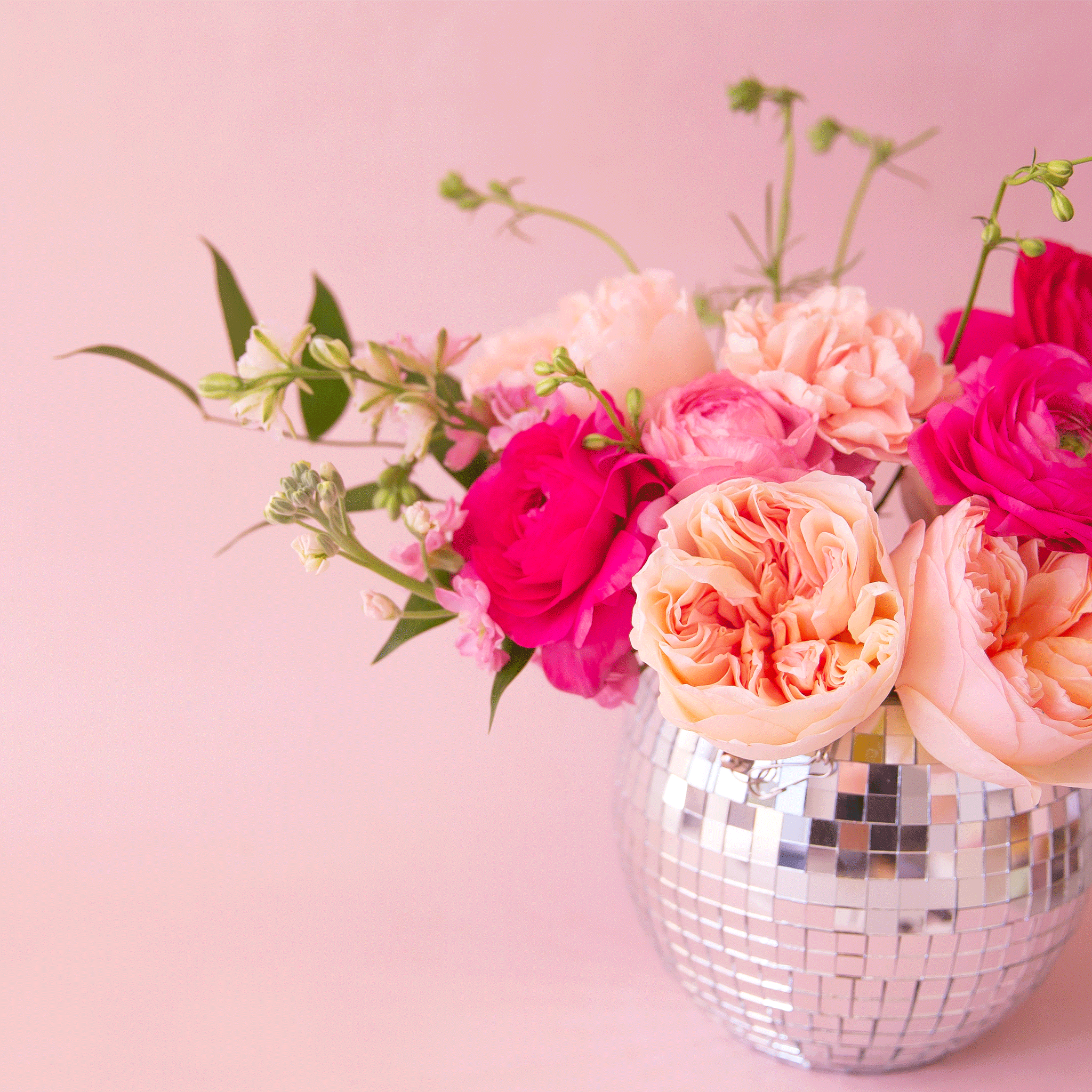 On a light pink background is a silver hanging planter with a pink flower arrangement in it.
