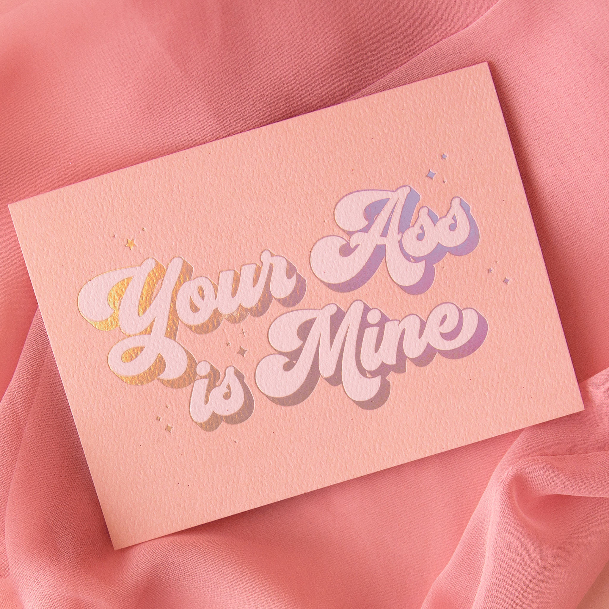 On a pink background is a peachy pink card with holographic and ivory text that reads, "Your Ass is Mine". 