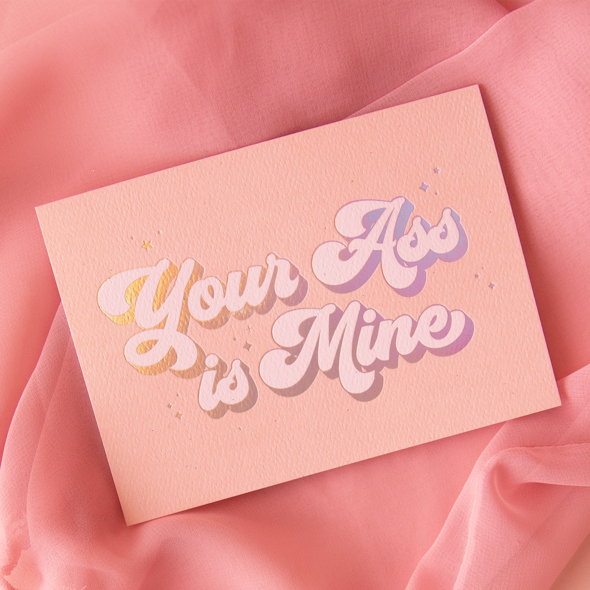 On a pink background is a peachy pink card with holographic and ivory text that reads, "Your Ass is Mine".