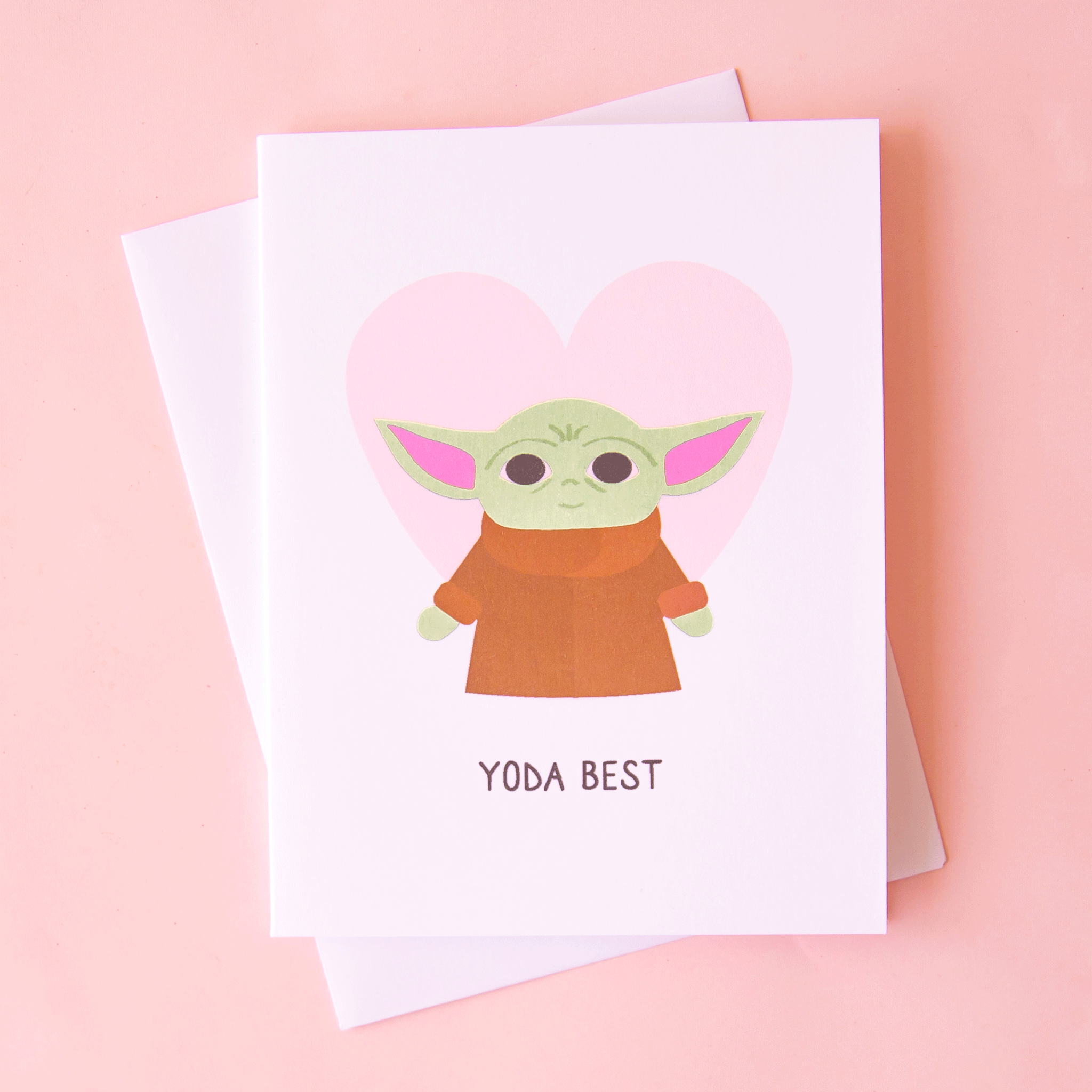 A white card with a light pink heart and a Yoda illustration in front along with black text that reads, "Yoda Best" along with a white envelope.