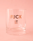 On a pink background is a clear glass mug with gold text across the front that reads, "Fuck It".