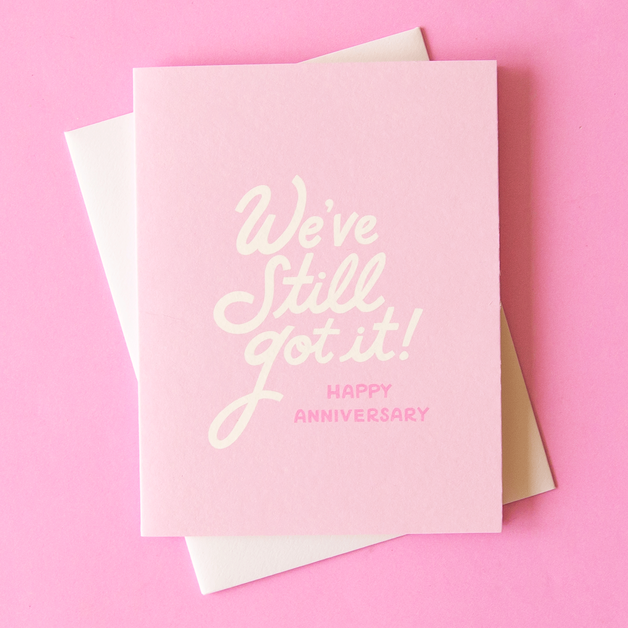 On a pink background is a light pink card with white text that reads, &quot;We&#39;ve Still Got It! Happy Anniversary&quot;.