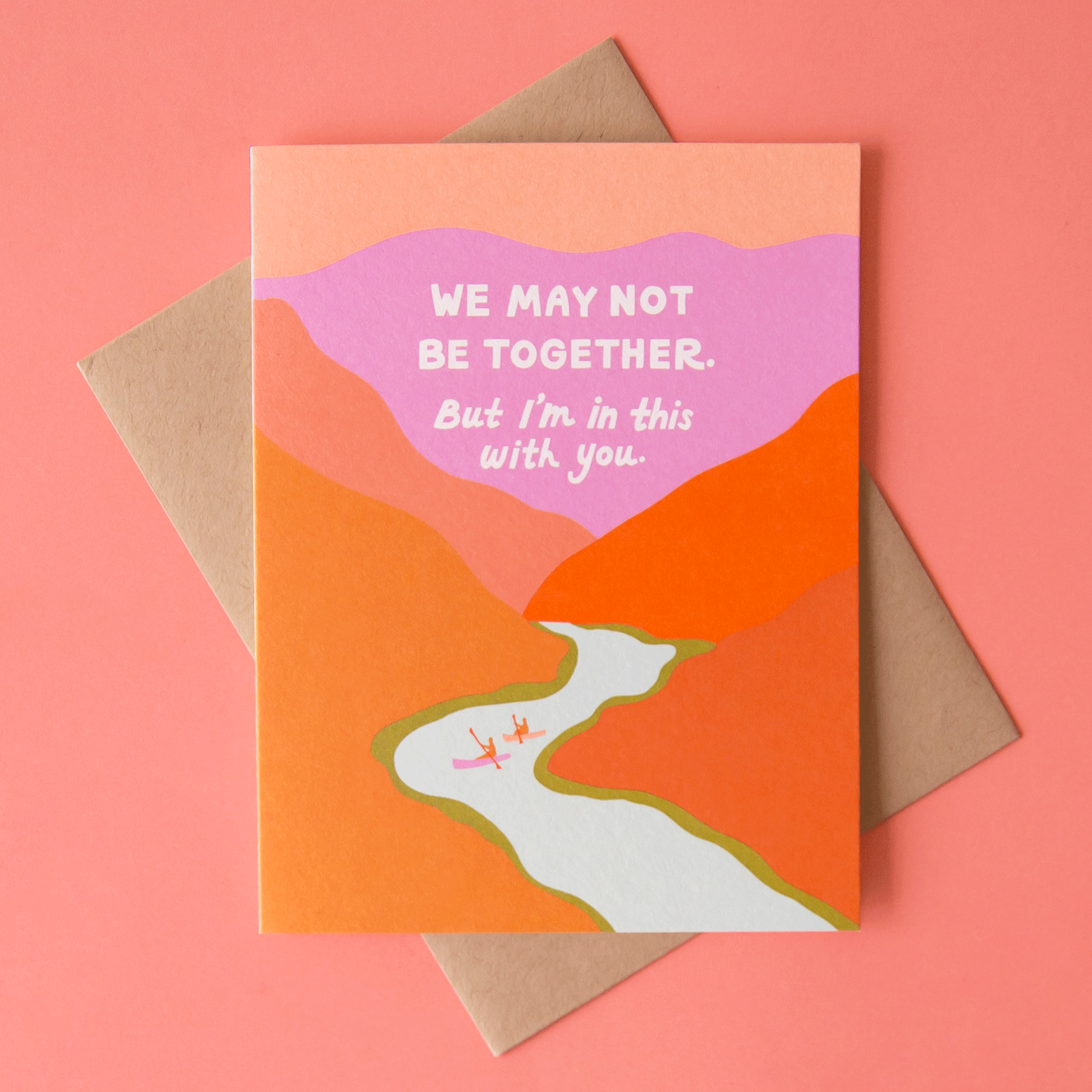 On a coral background is a vibrant card with shades of orange, red, pink and purple making up a mountainous range with a river running through it along with white text at the top of the card that reads, &quot;We May Not Be Together But I&#39;m In This With You&quot;.