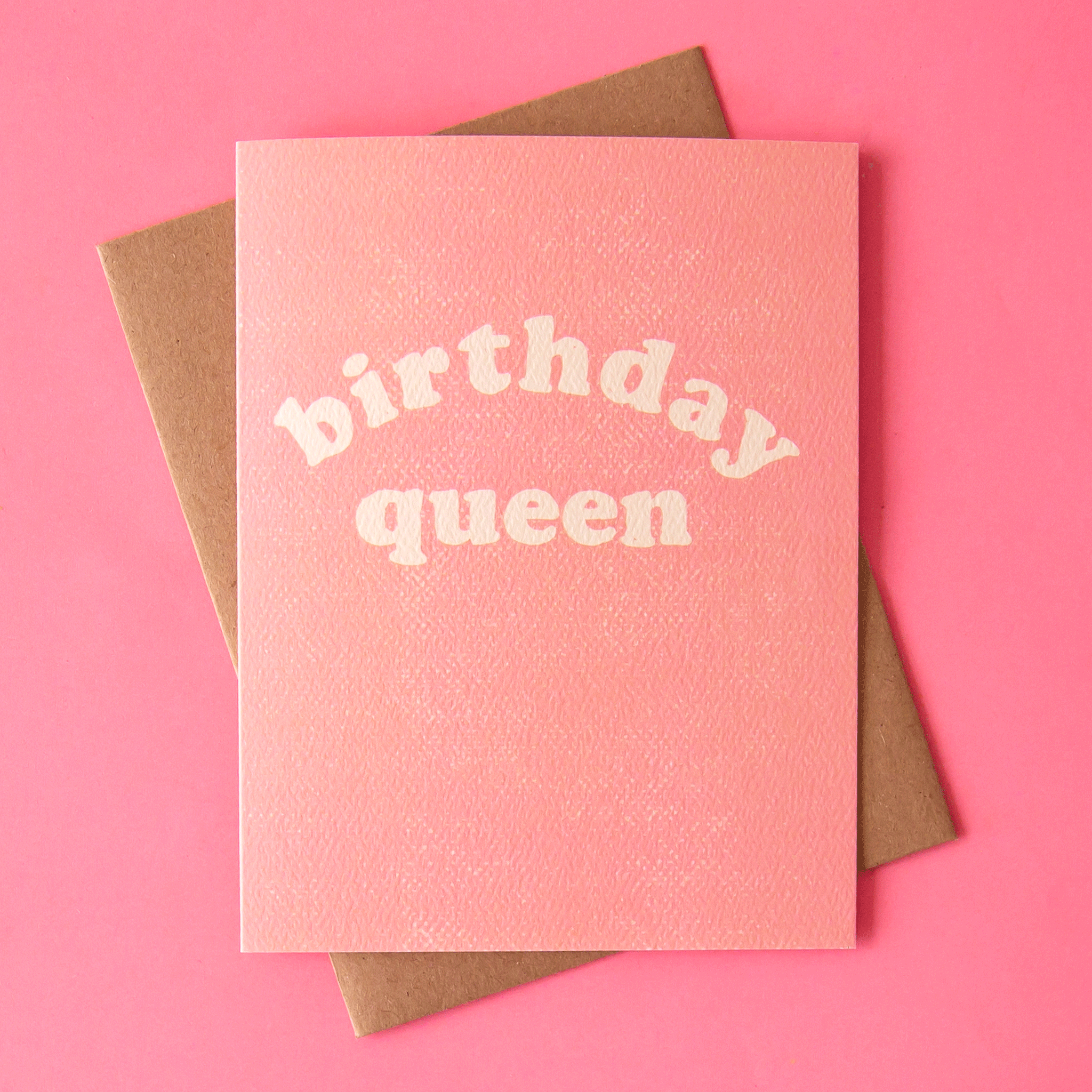 On a pink background is a pink birthday card with a Kraft brown envelope that says, "birthday queen" in white text.