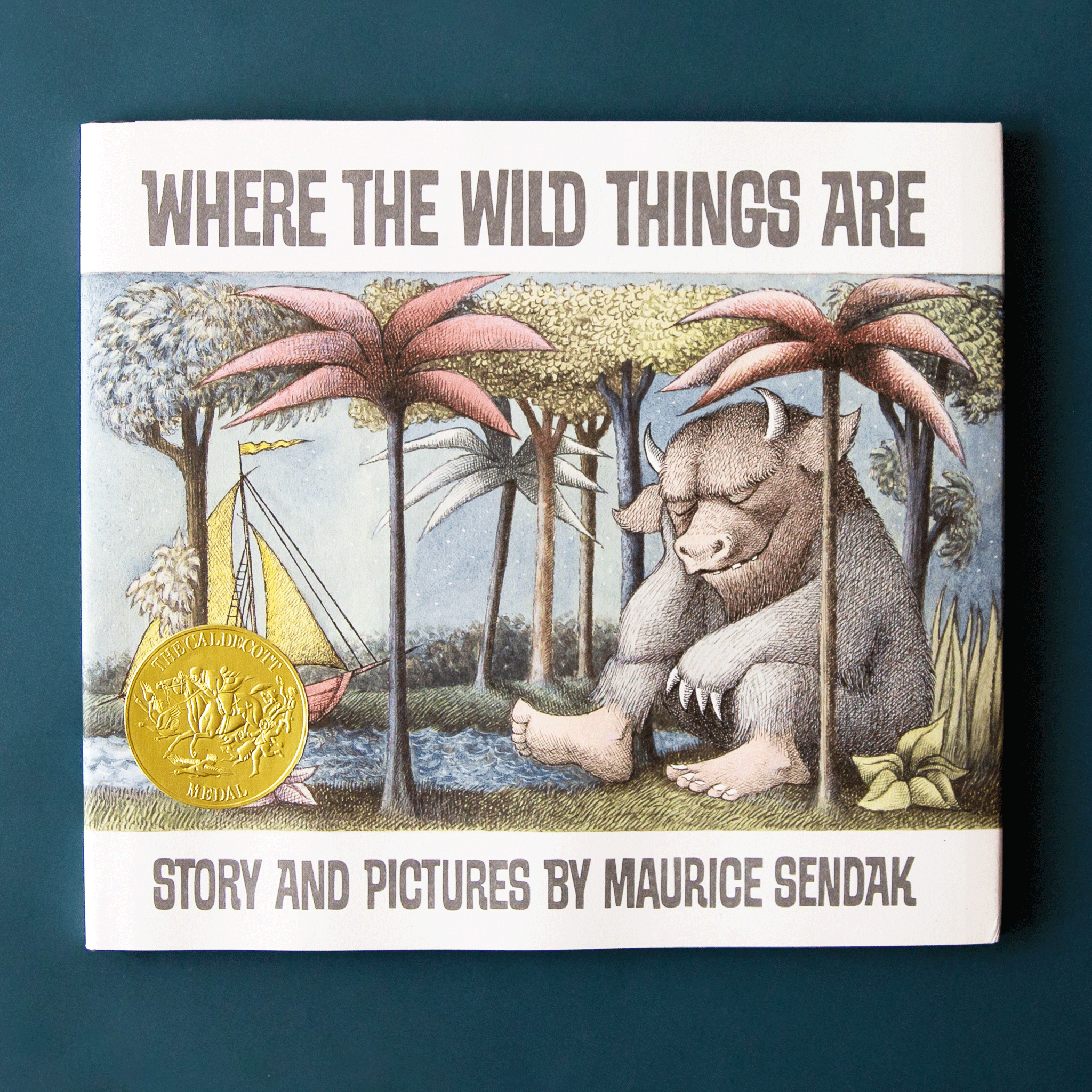 This classic children's story book features a dreamy forest homing a large mystic creature with pale blue fur, sharpened claws and round horns. The creature sits peacefully among the trees and across from a pond filled with a soft yellow and red sail boat. The book is titled 'Where the Wild Things Are' in pale teal capital lettering above.