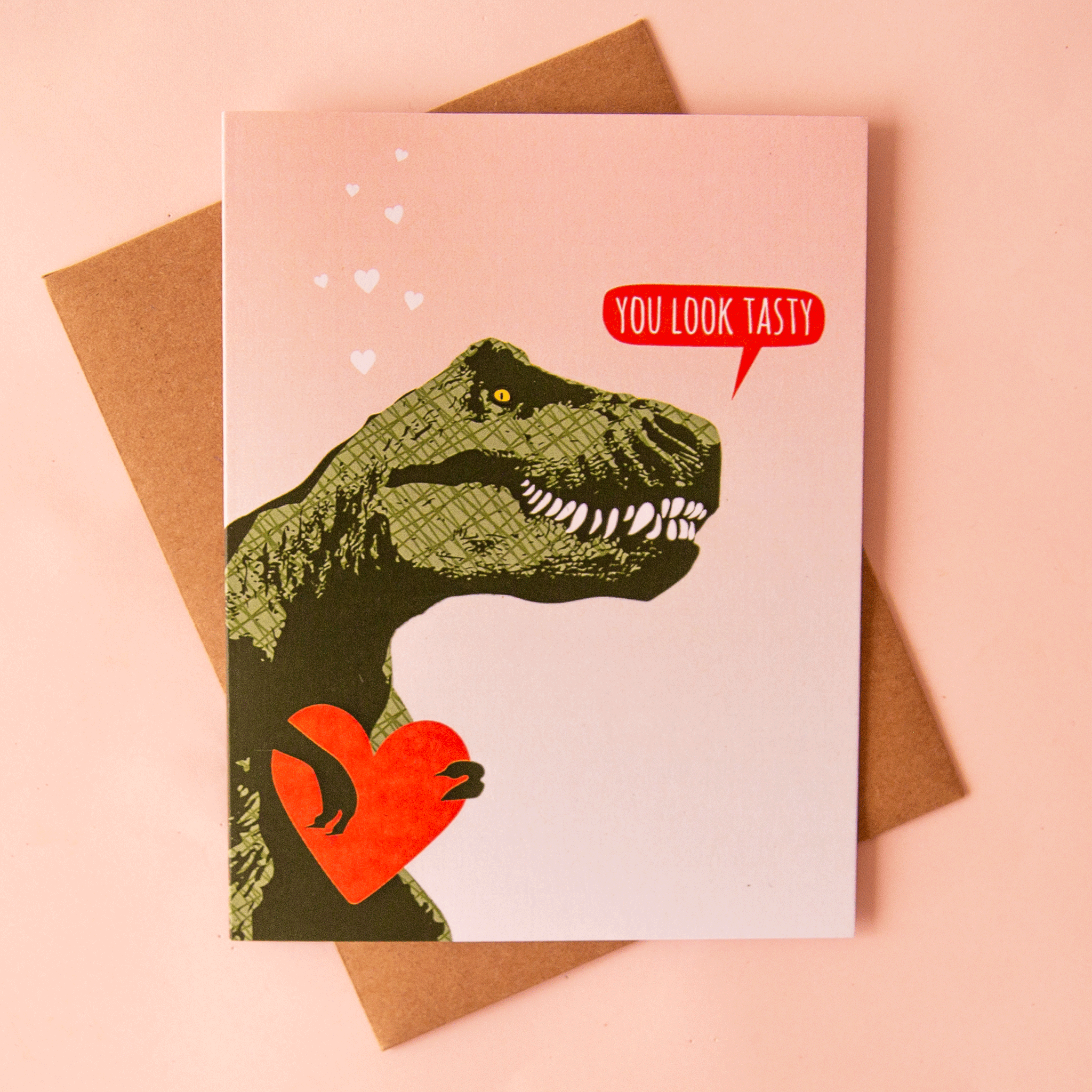 On a pink background is a pink card with a green t-rex illustration that's holding a heart and a speech bubble that reads, "You Look Tasty".
