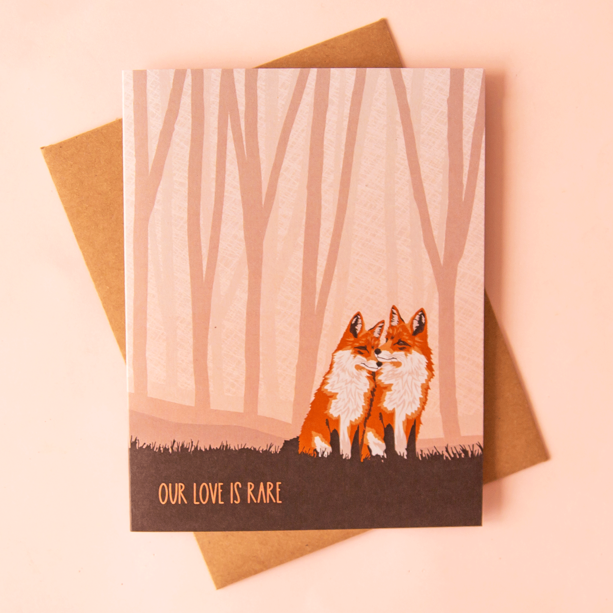 On a pink background is a tan card with a woodland background that has two orange foxes cuddling and text along the bottom that reads, "Our Love Is Rare". 