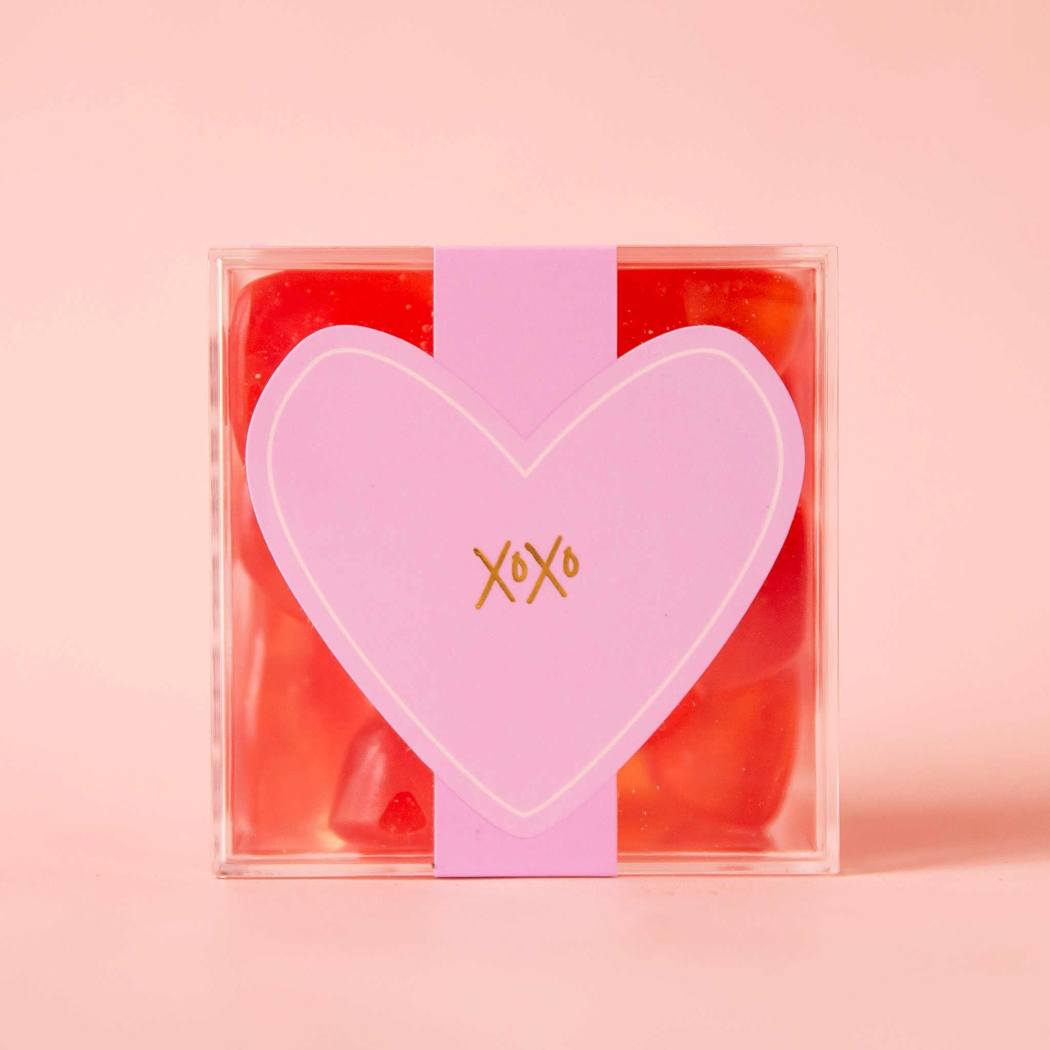 On a pink background is an acrylic clear box of red heart shaped gummy candies.