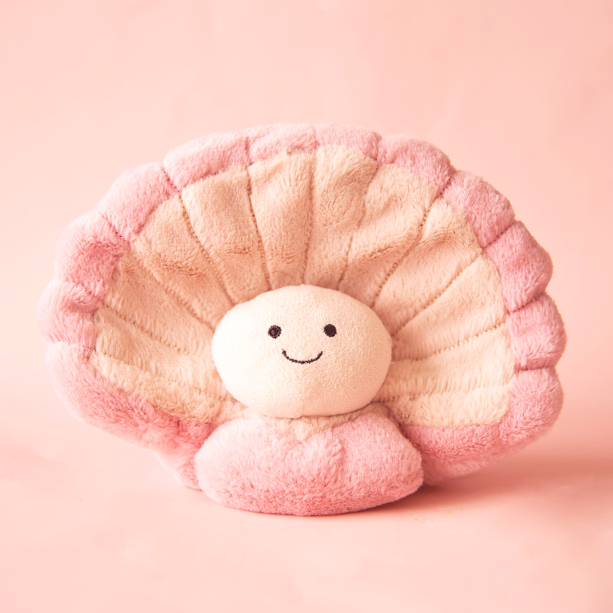 On a pink background is a clam shaped stuffed toy with a smiling cream colored pearl nestled inside of the pink and tan clam shaped shell. 