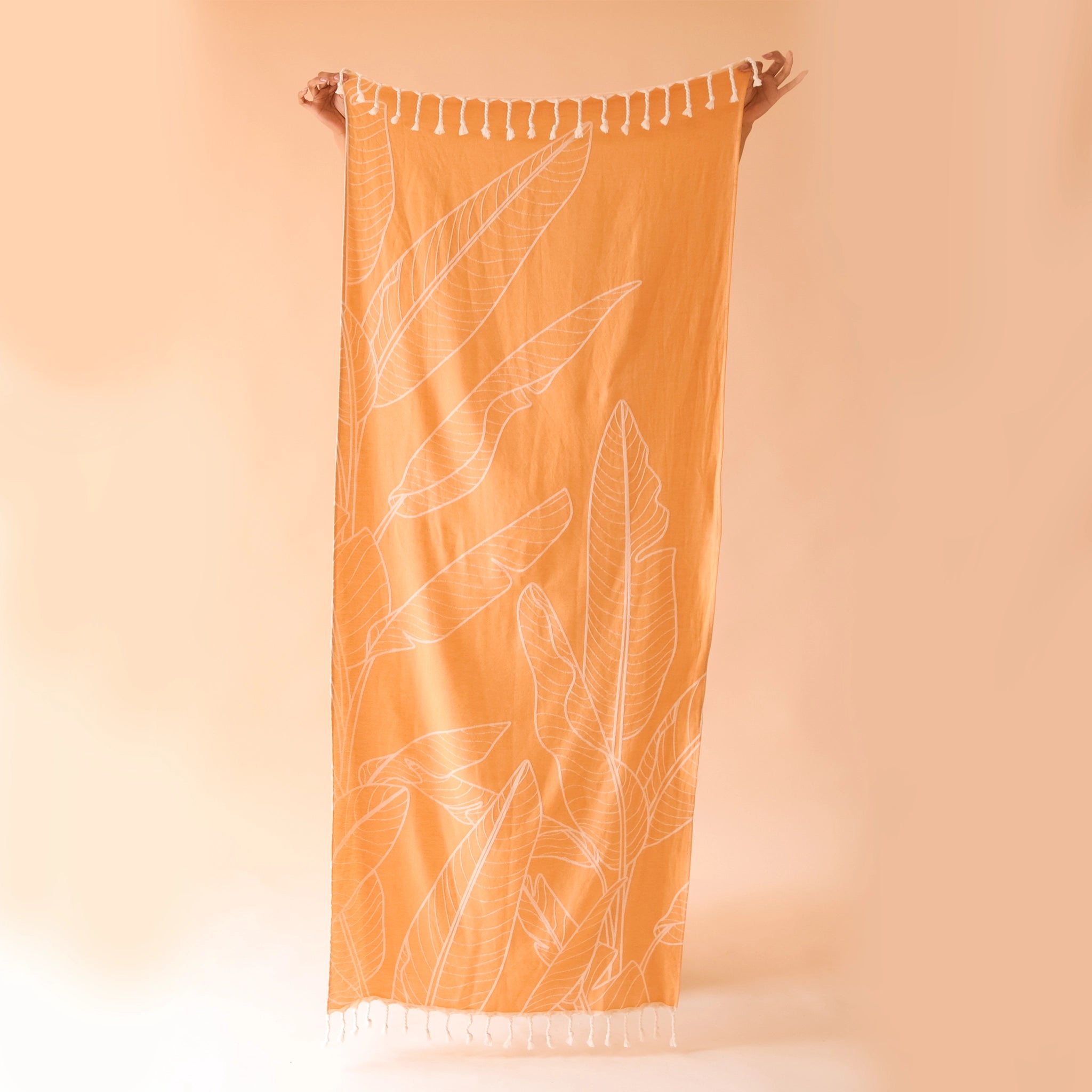 An orange beach towel with a banana leaf print and white tassels on each end.