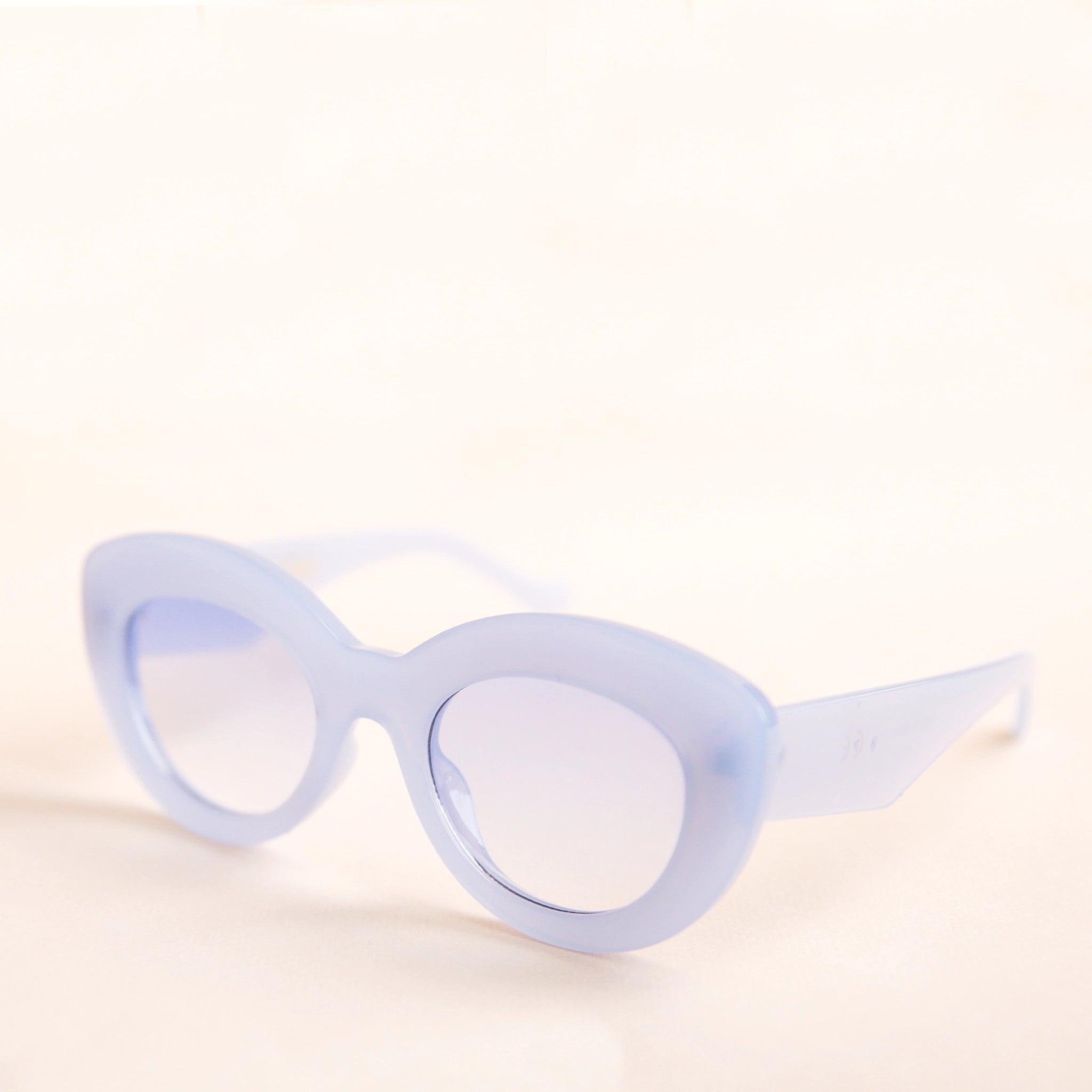 On a neutral background is the Gemma Sunglasses in a light blue shade. They have a rounded shape with a slight cateye flair at the corners. The frame material is a durable plastic and the lenses are a similar tone.