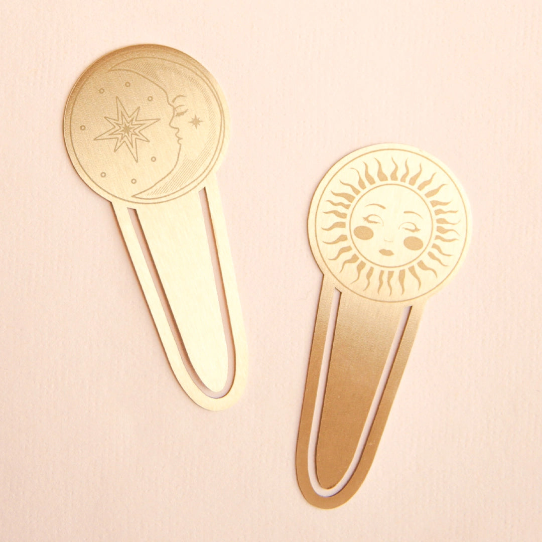 On a pink background is two gold metal page tabs with a sun and moon design on each. 
