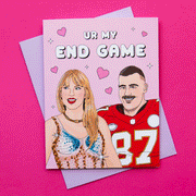 On a hot pink background is a pink card with an illustration that looks like Taylor Swift and Travis Kelce along with text above them that reads, "Ur My End Game". The card comes with a light purple envelope.