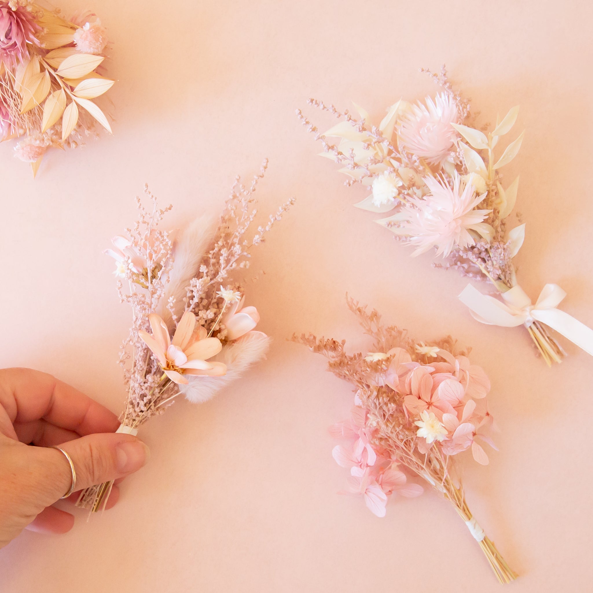 A hand holds a small dried flower bouquet while three similar pastel pink and cream bouquets are arranged on a soft peach background.