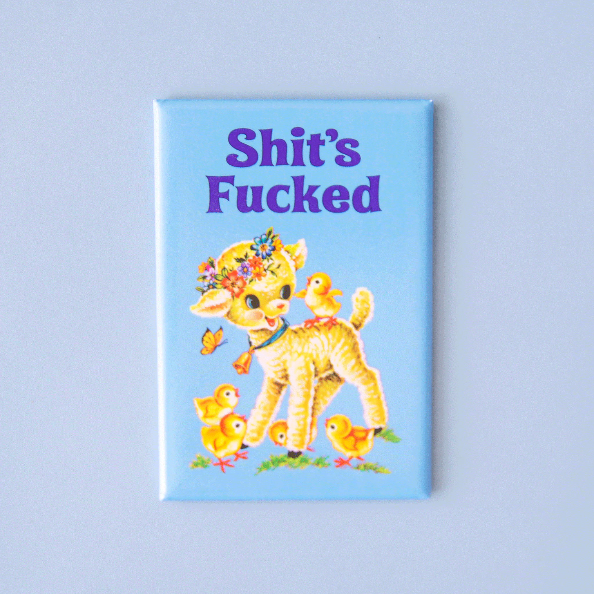 On a blue background is a blue rectangle magnet with a graphic of a yellow lamb and baby chicks along with blue text above that reads, "Shit's Fucked". 