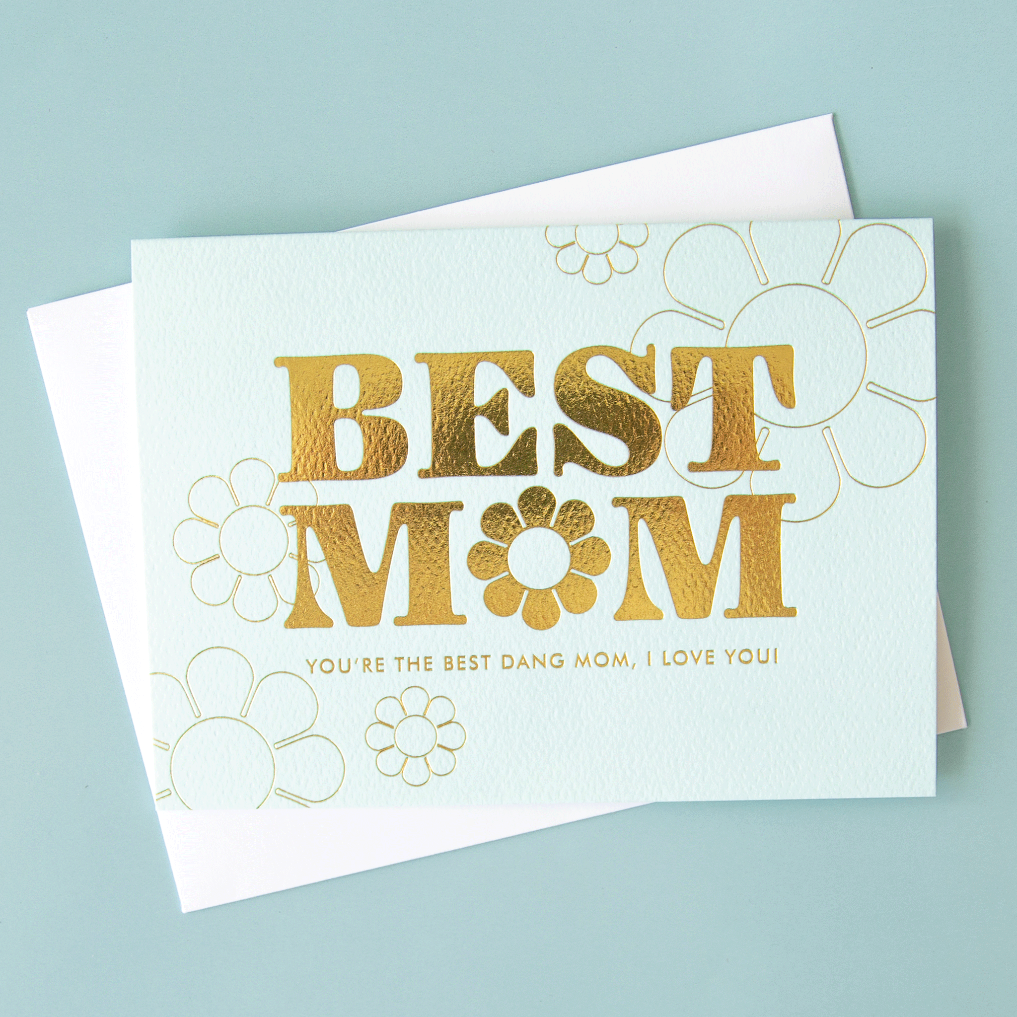On a blue background is a light blue card with gold foiled text that reads, &quot;Best Mom You&#39;re The Best Dang Mom, I Love You&quot; along with a flower shape in the place of the &#39;O&#39; on &#39;Mom&#39;. 