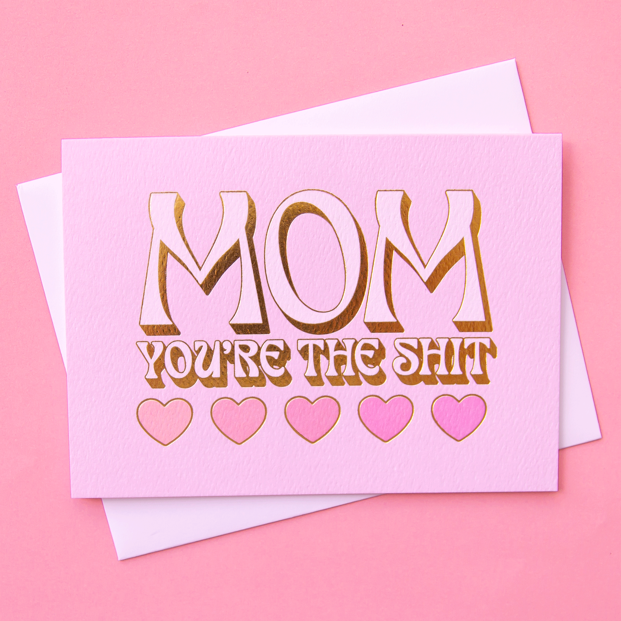 On a pink background is a pink card with heart shapes and text above that reads, &quot;Mom You&#39;re The Shit&quot;.