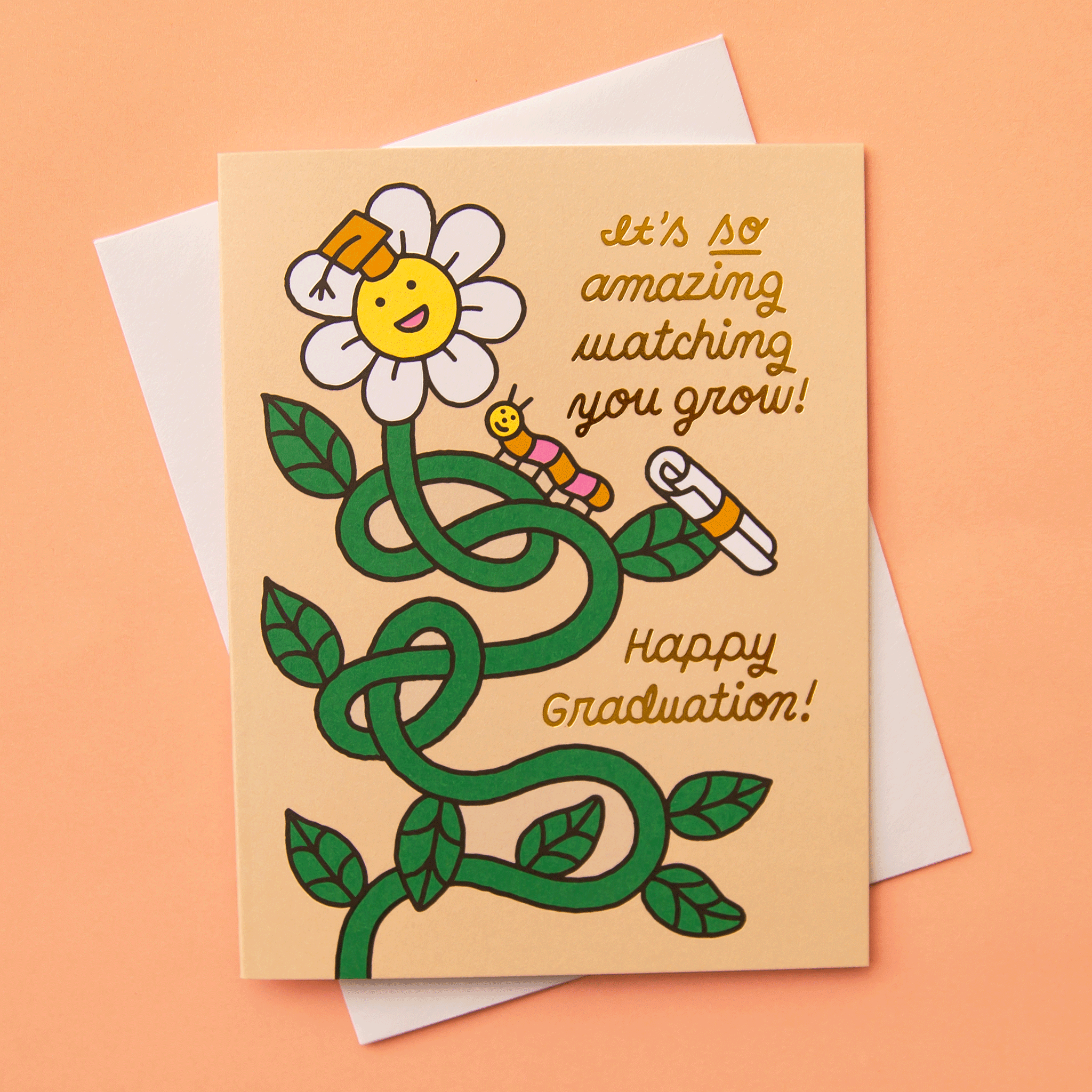 On a peach background is a light tan card with a daisy graphic with a graduation cap on and gold text that reads, "It's so amazing watching you grow! Happy Graduation!". 