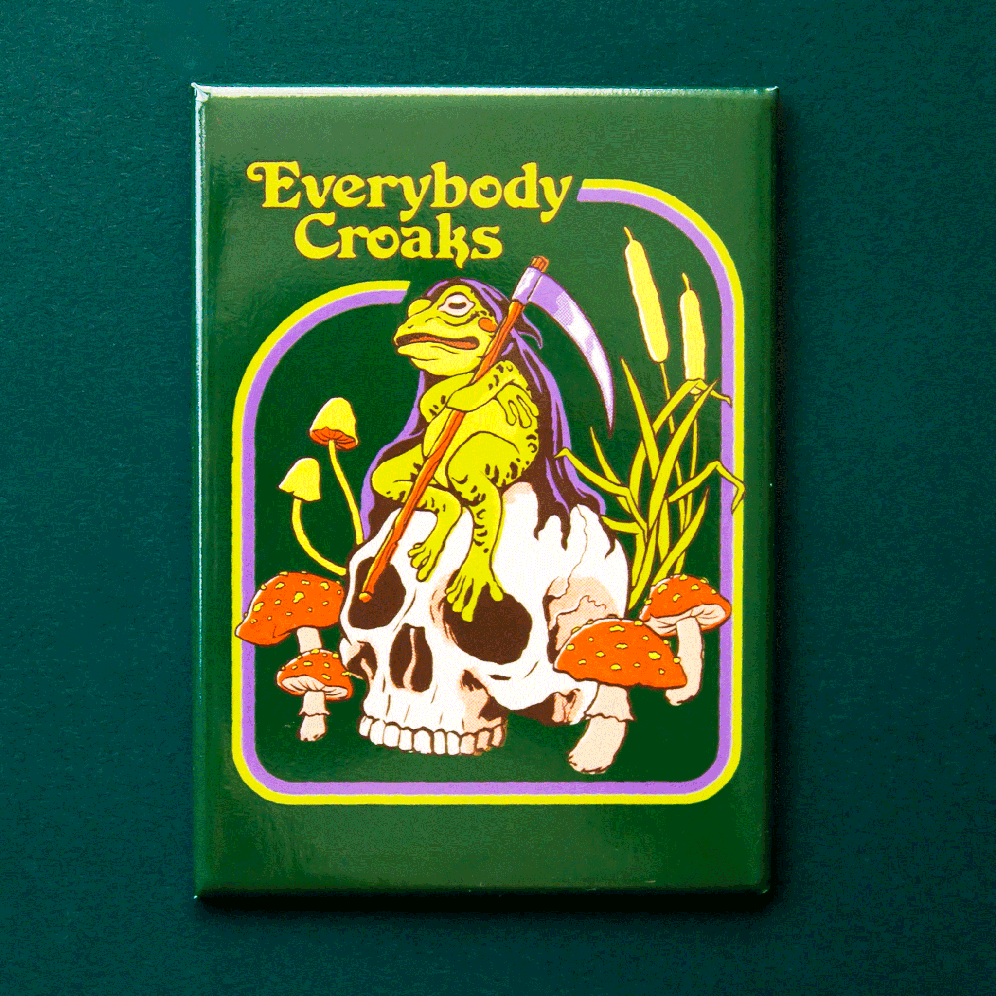 On a green background is a green rectangle magnet with a graphic of a toad sitting on a skull with text above that reads, &quot;Everybody Croaks&quot;. 