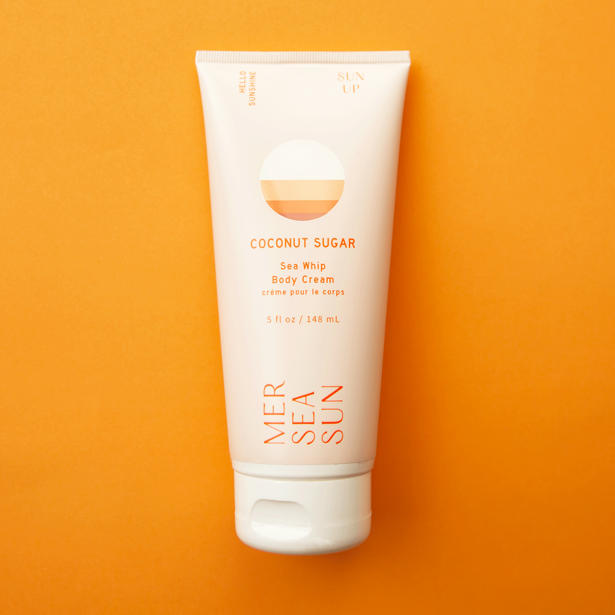 On an orange background is a light pink bottle of lotion with orange text on the front that reads, "Mersea Sun Coconut Sugar Sea Whip Body Cream".