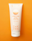 On an orange background is a light pink bottle of lotion with orange text on the front that reads, "Mersea Sun Coconut Sugar Sea Whip Body Cream".