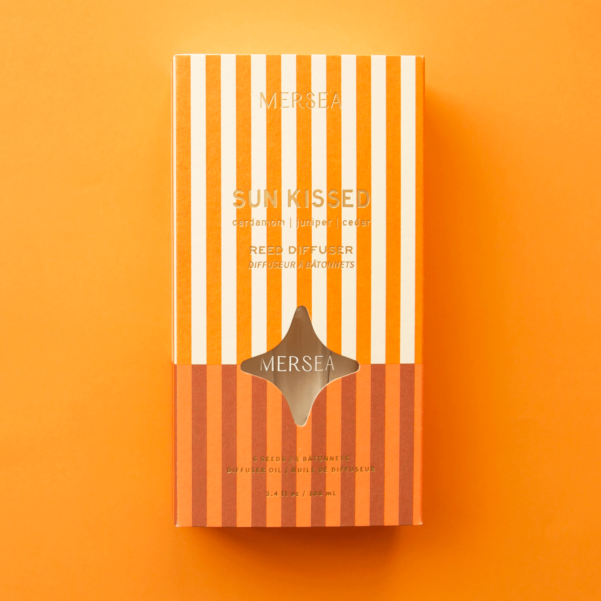 On an orange background is an orange and white striped box filled with a reed diffuser. 