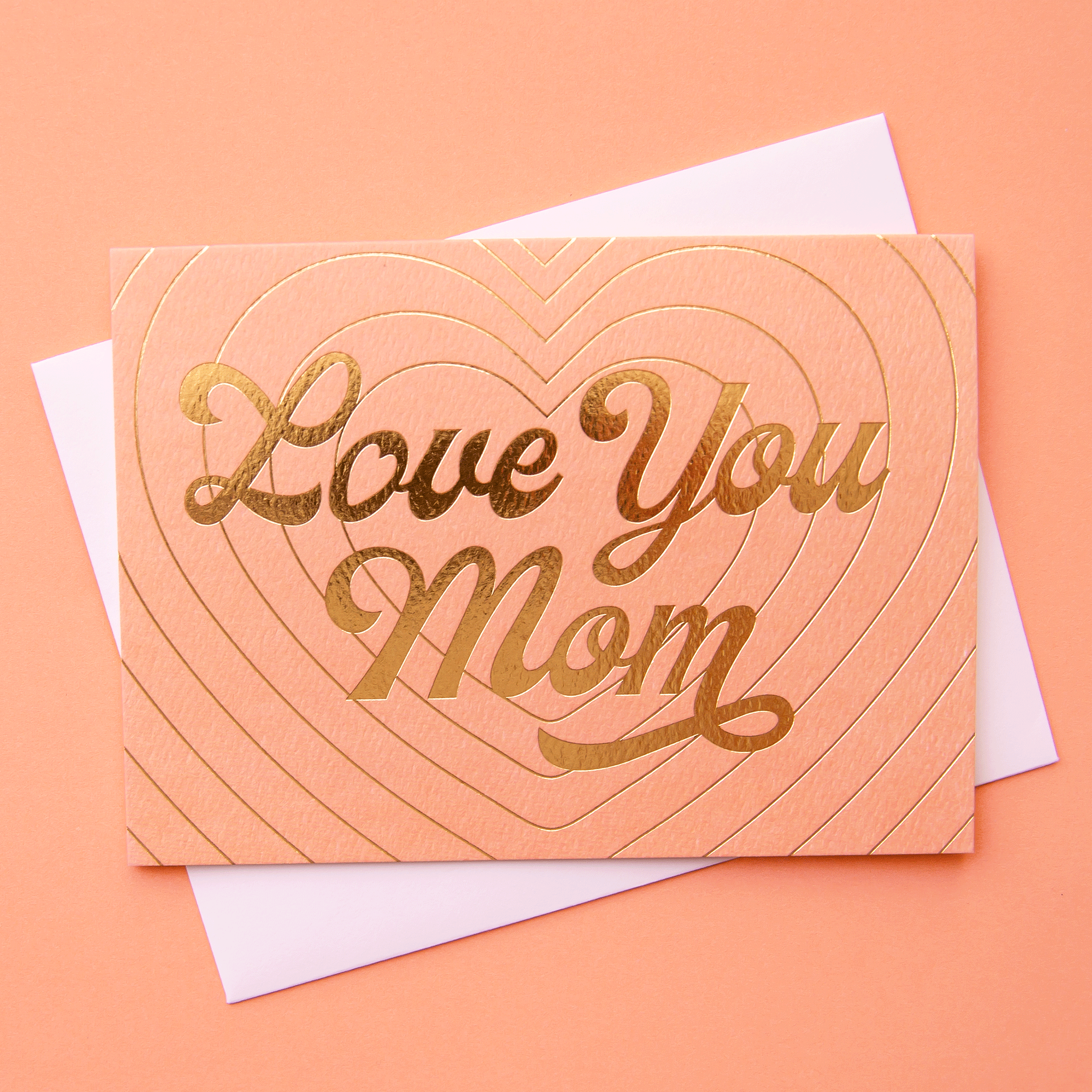 Peach card that reads &#39;love you mom&#39; in gold foil. The card is filled with layers of a beaming gold foil heart design behind and is accompanied by with a white envelope.