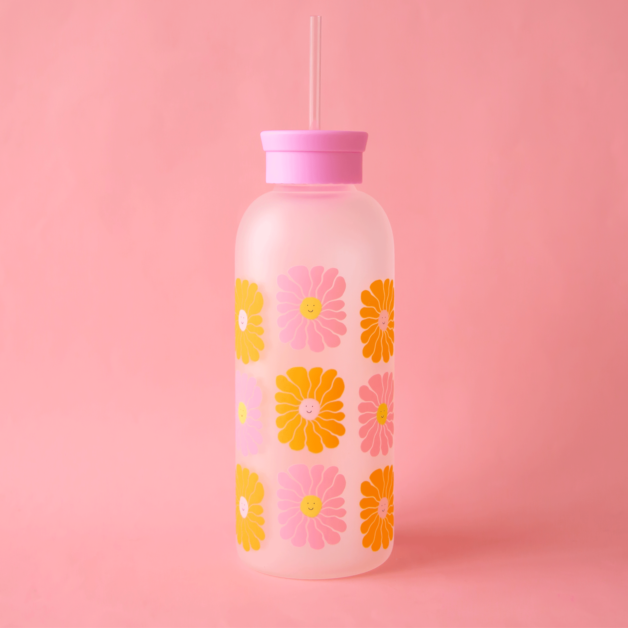 On a pink background is a water bottle with a silicone lid, glass straw and a wavy smiley face daisy print in pink and orange shades. 