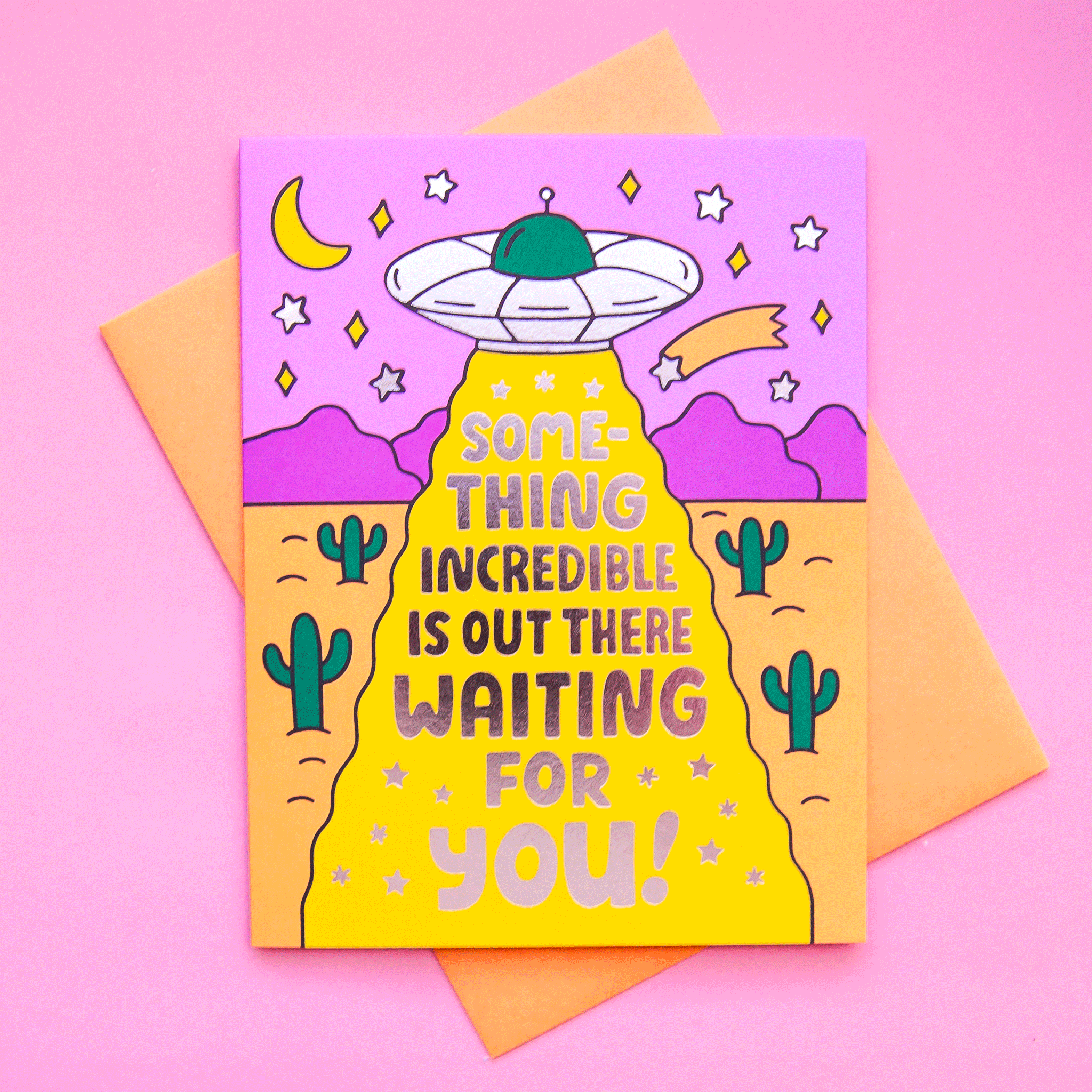 A vibrant greeting card with an illustration of a spaceship floating in a desert, detailed with twinkling stars, green cacti, and a fuchsia mountain scape and reads, "Something incredible is out there waiting for you!". Also included is a coordinating orange envelope.