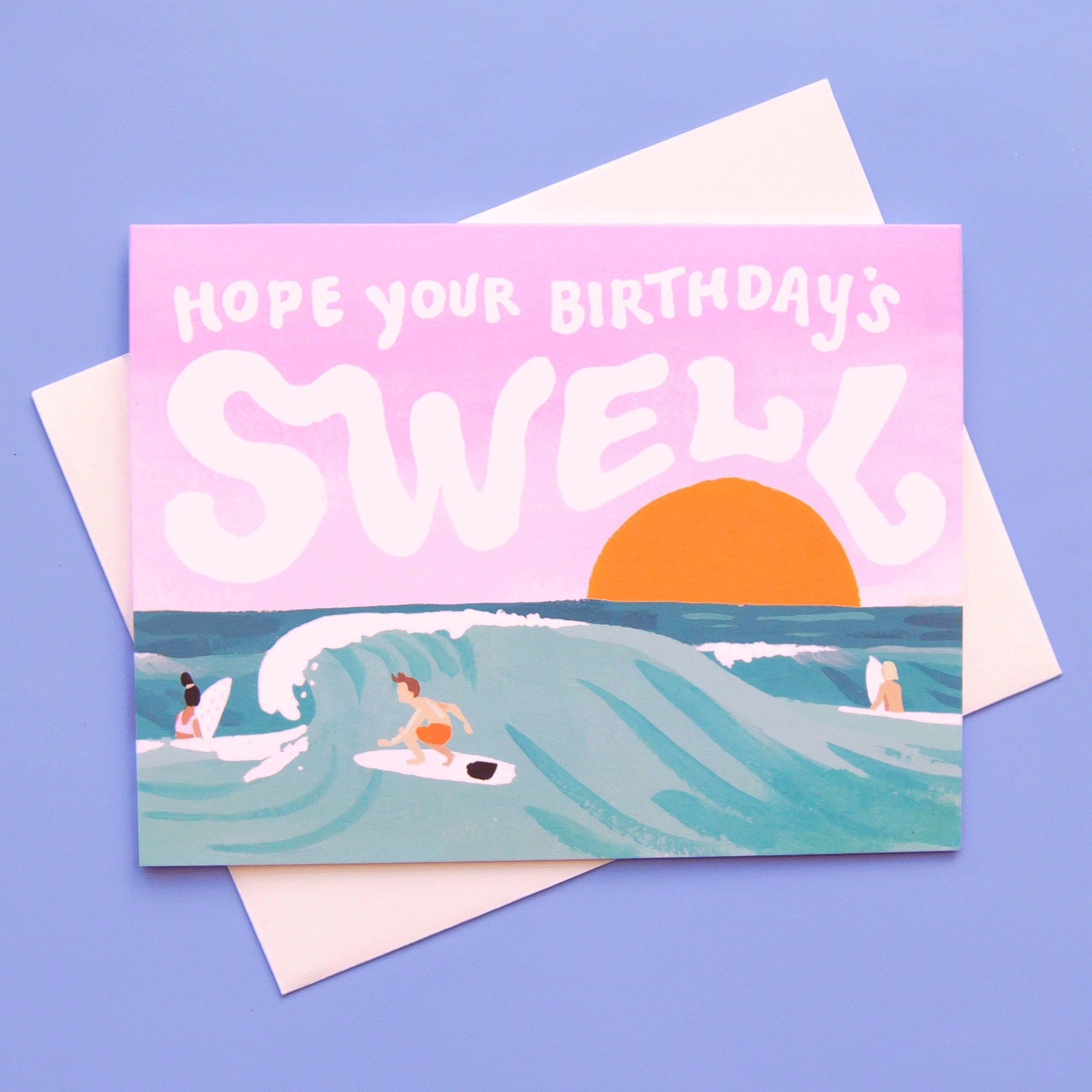 On top of a white envelope is a horizontal card. The card is a drawing of a blue ocean with a wave and a person surfing the wave. Behind the ocean is a purple sky with a yellow, setting sun. In the sky is white text that reads ‘hope your birthday’s swell.’