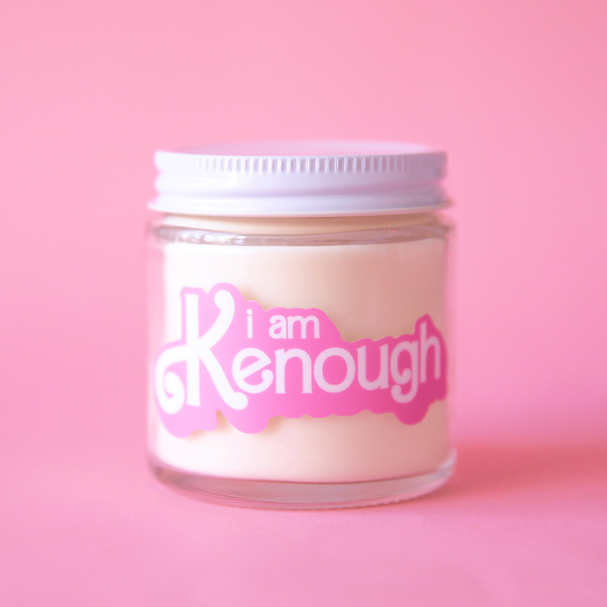 On a pink background is a glass candle with a white lid and pink text on the front that reads, &quot;I am Kenough&quot;.