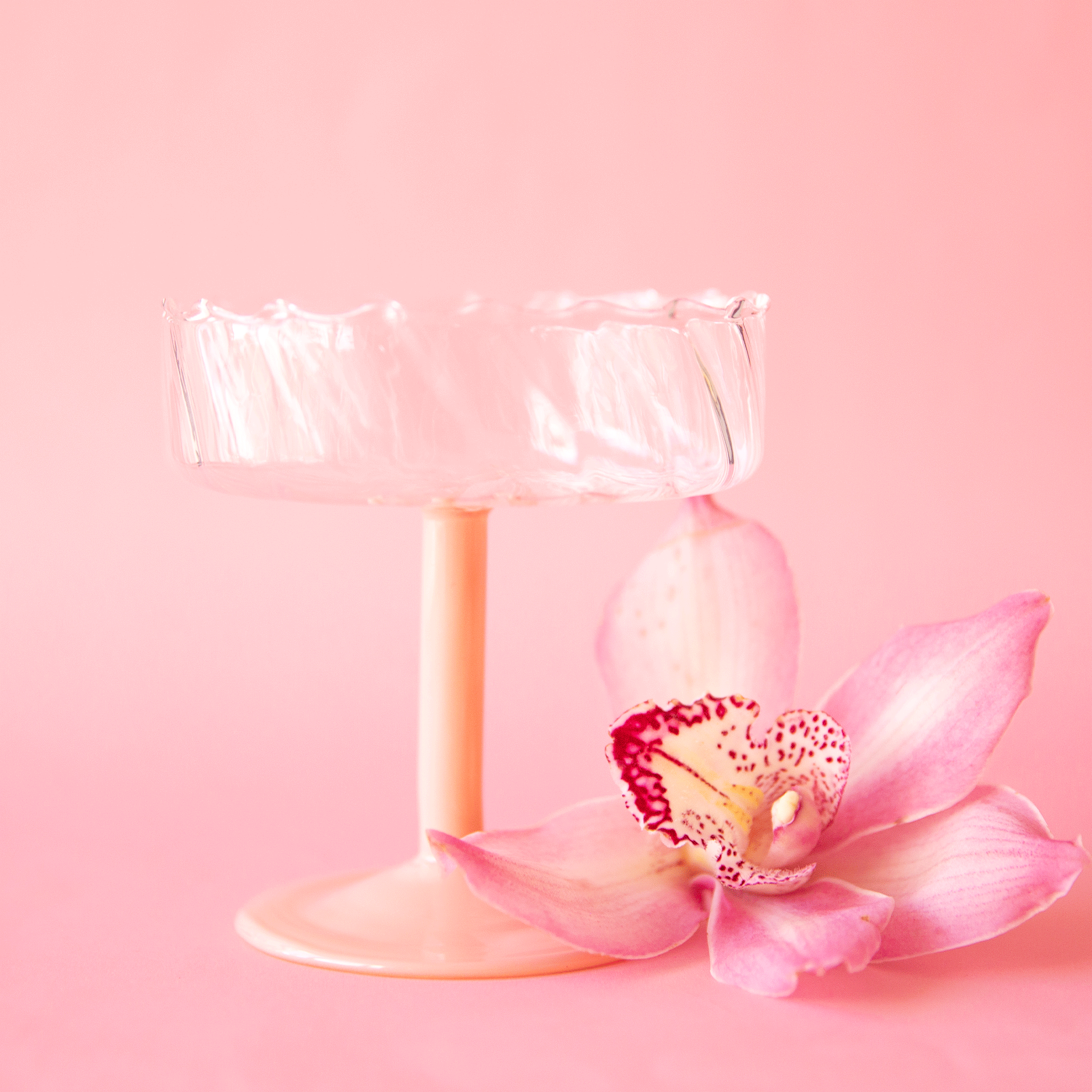 On a pink background is a wavy glass coupe with a pink stem. 