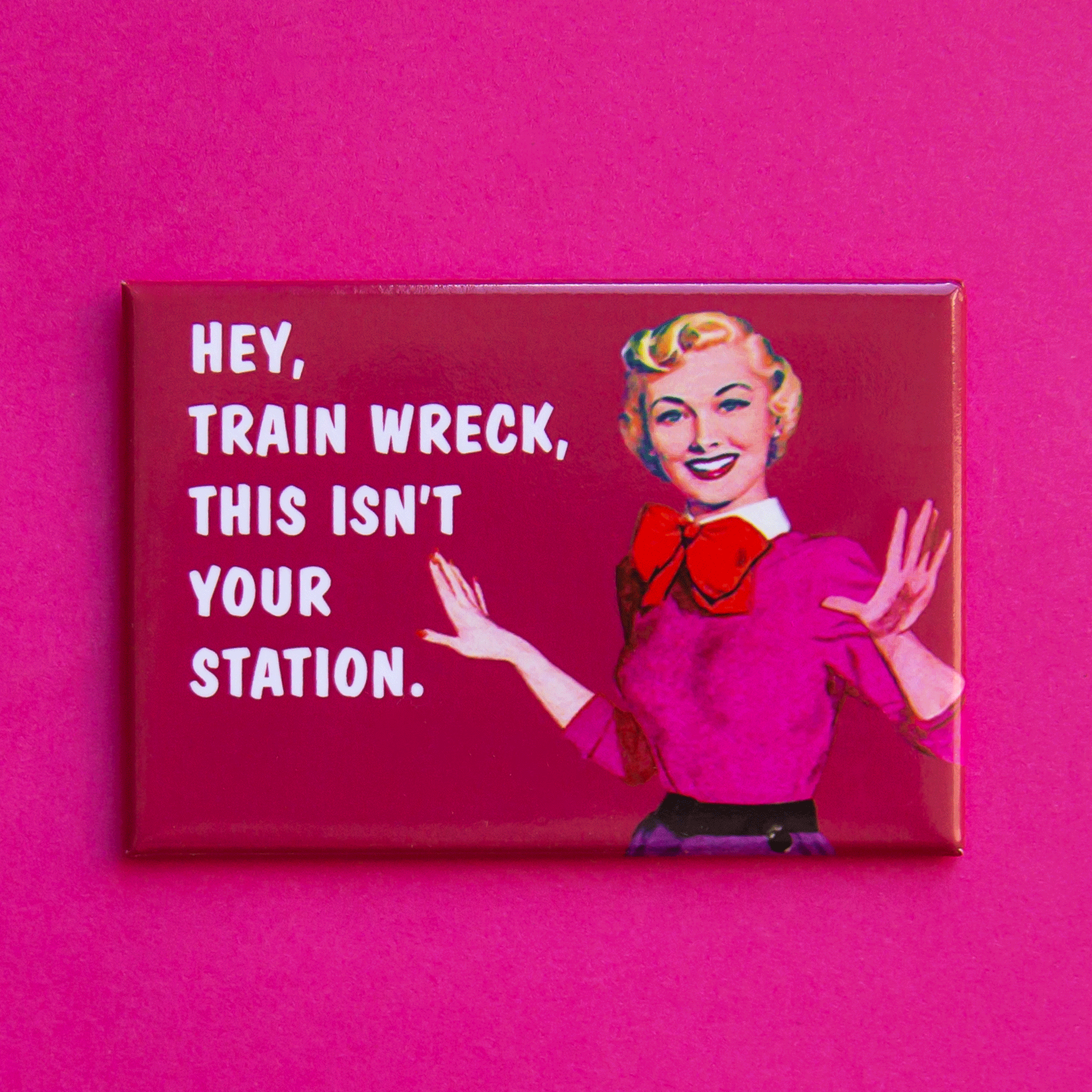 On a pink background is a pink magnet with a vintage graphic of a woman in a pink sweater and white text that reads, "Hey, Train Wreck This Isn't Your Station.". 
