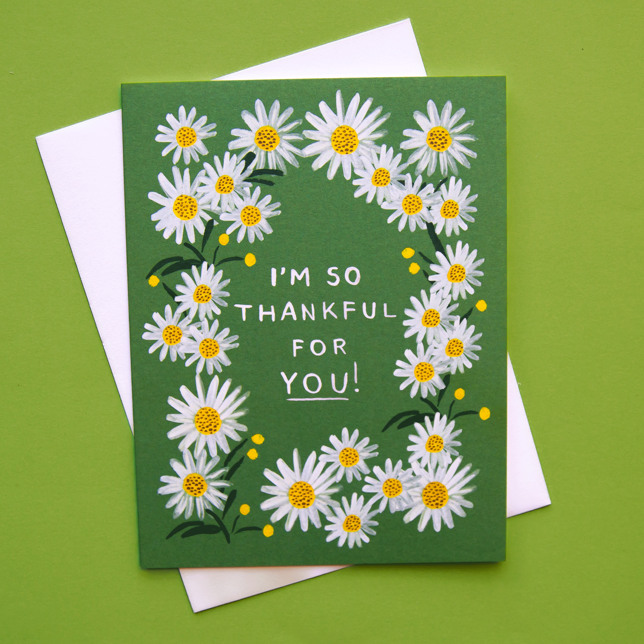 A photograph of a green card and a white envelope behind it. The card has a white daisy border around white text in the center that reads, &quot;I&#39;m So Thankful For You!&quot;.
