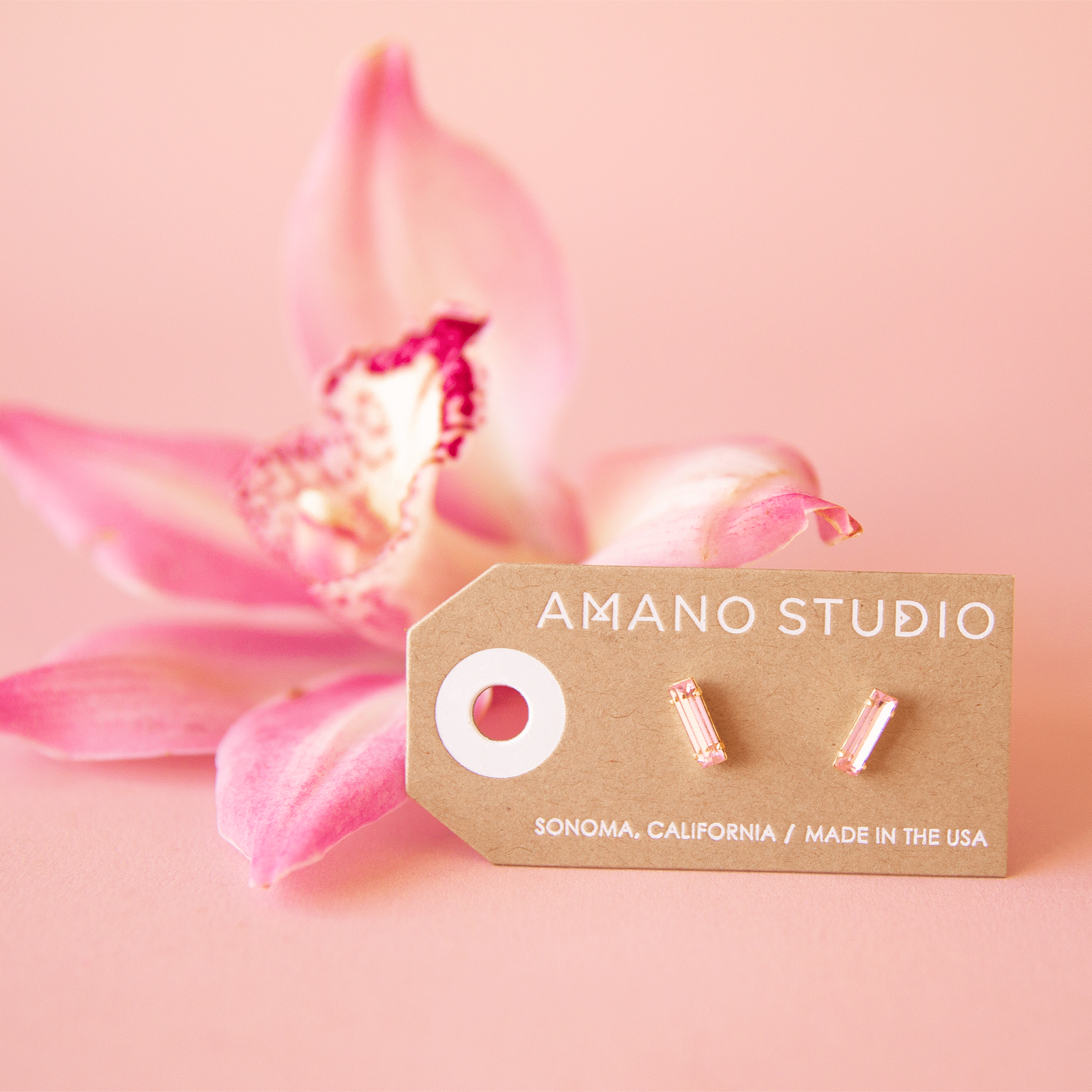 On a pink background is a gold plated of pair of baguette stud earrings with a pink stone in the center.